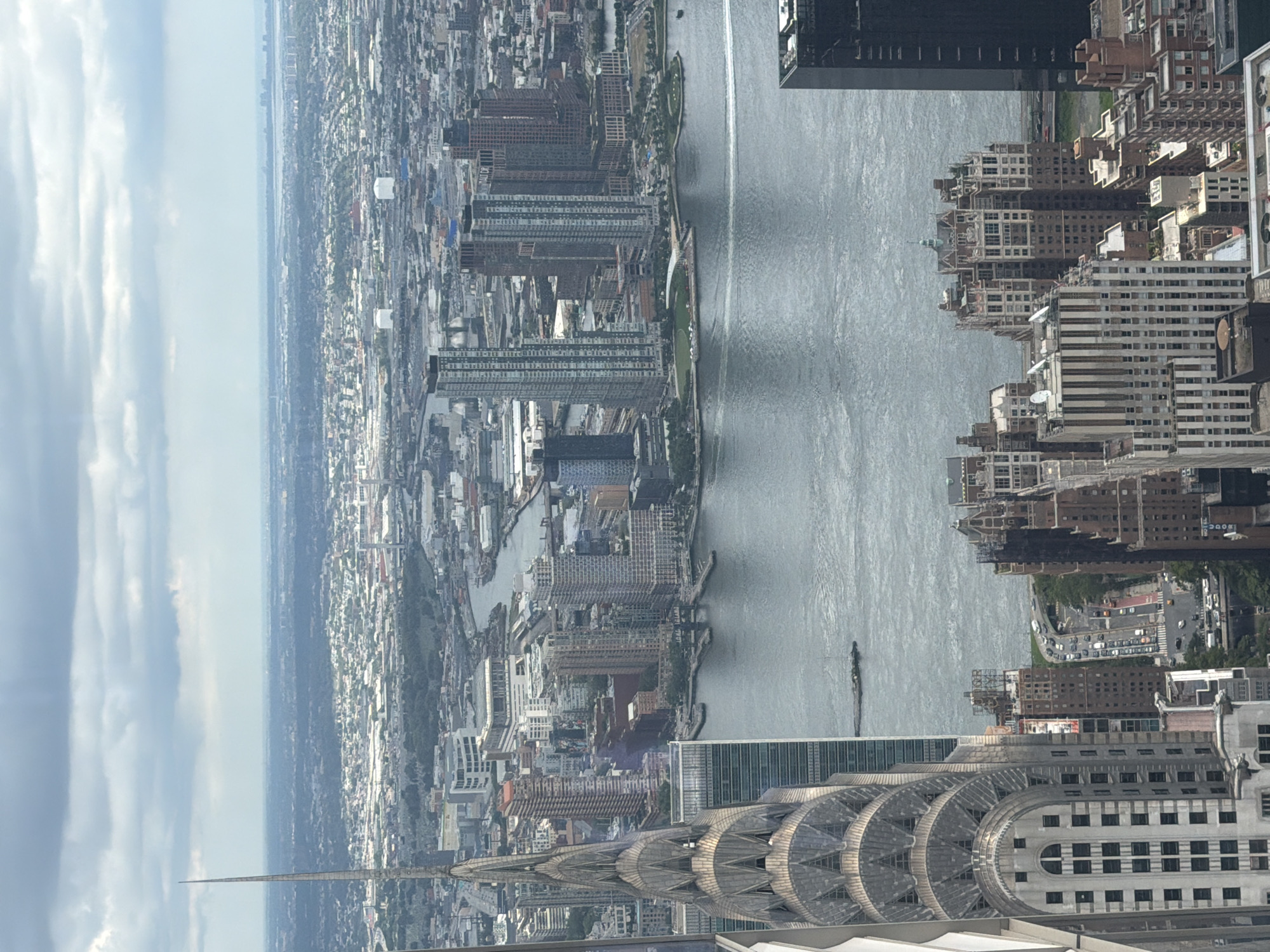 This is an aerial view of New York City. In the foreground, the iconic Chrysler Building is visible on the left, with its distinctive art deco spire. The East River flows between Manhattan and Queens. Numerous skyscrapers populate the Manhattan skyline, including the long, slender buildings on the right bank of the river, which appear to be in Long Island City, Queens. The distant background shows the hazy expanse of other boroughs of New York City and possibly New Jersey. The sky is overcast with clouds. No people are discernable in the image. The scene captures a dense urban landscape with significant architectural landmarks.