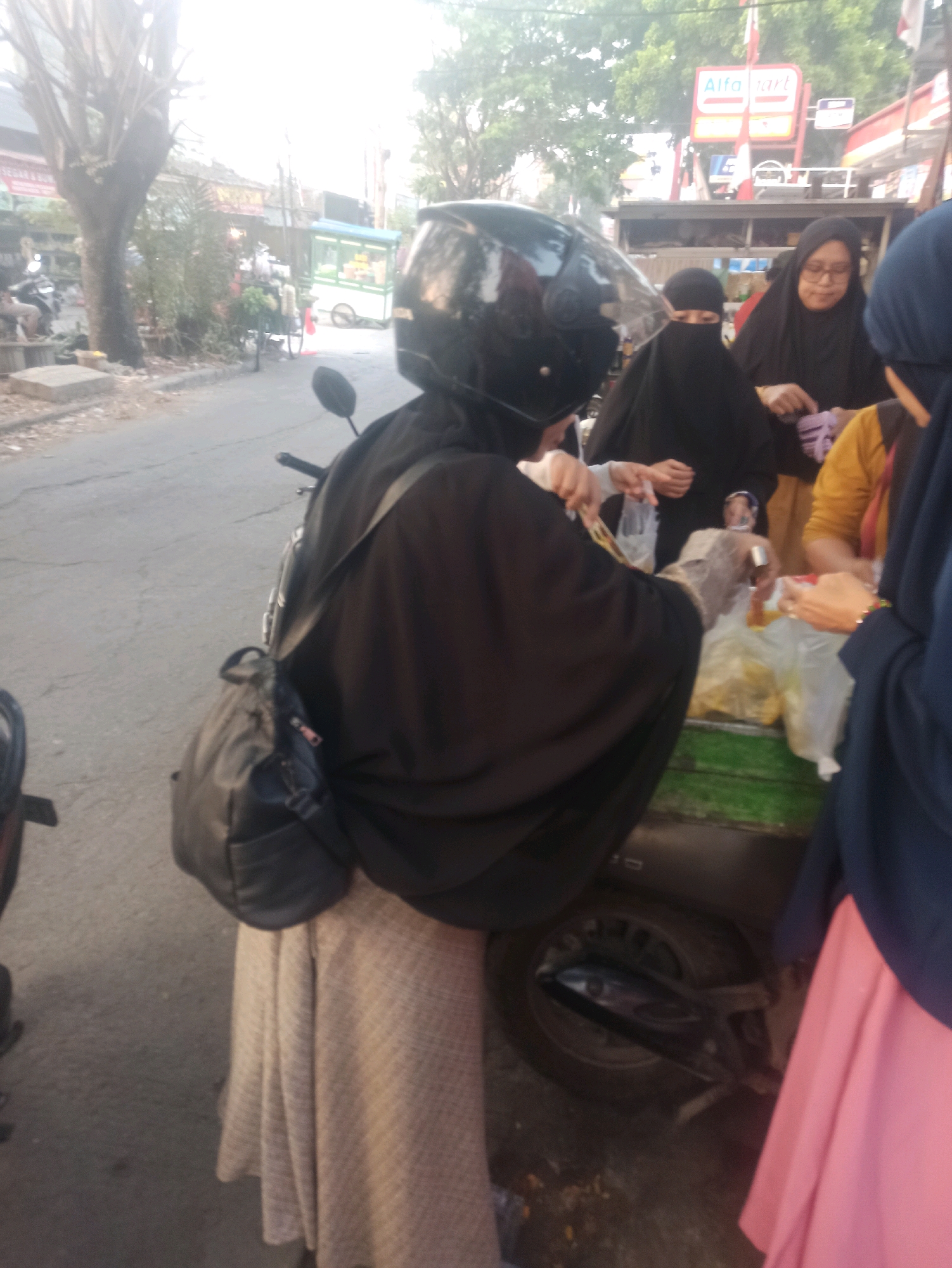 In Telagamurni, Indonesia, a group of women are seen at a roadside stall. One woman, wearing a black hijab and a beige checkered skirt, is seated on a motorcycle and appears to be in the process of purchasing food items from a vendor. She is wearing a helmet and has a black bag slung over her shoulder. The vendor, also a woman wearing a black hijab, is handing plastic bags containing food to another woman, who is wearing a yellow top and a dark blue hijab. Behind them, another woman in a dark blue hijab is visible. The stall itself is green and white. The background shows a street with trees and a building with a red and white sign that reads "Alfa Mart". The overall scene suggests a typical marketplace environment.