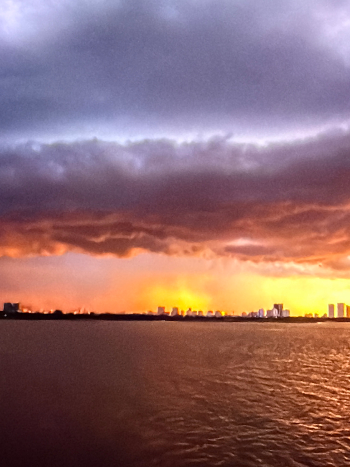 A wide-angle, low-resolution, over-saturated photograph captures a vibrant sunset over a body of water, possibly the Mediterranean Sea, in front of a distant cityscape. The sky is dominated by dramatic clouds, with the upper portion rendered in shades of lavender and dark gray, transitioning to intense oranges and yellows near the horizon. The cityscape, situated on the distant shore, is a blurred silhouette of buildings. The water in the foreground reflects the warm hues of the sunset, creating a textured, shimmering surface. The overall impression is one of natural beauty and urban development coexisting. The photograph does not contain any identifiable people or specific actions. The location is not explicitly identifiable as Kafr Yasif, Israel, based solely on the visual information provided in the image.