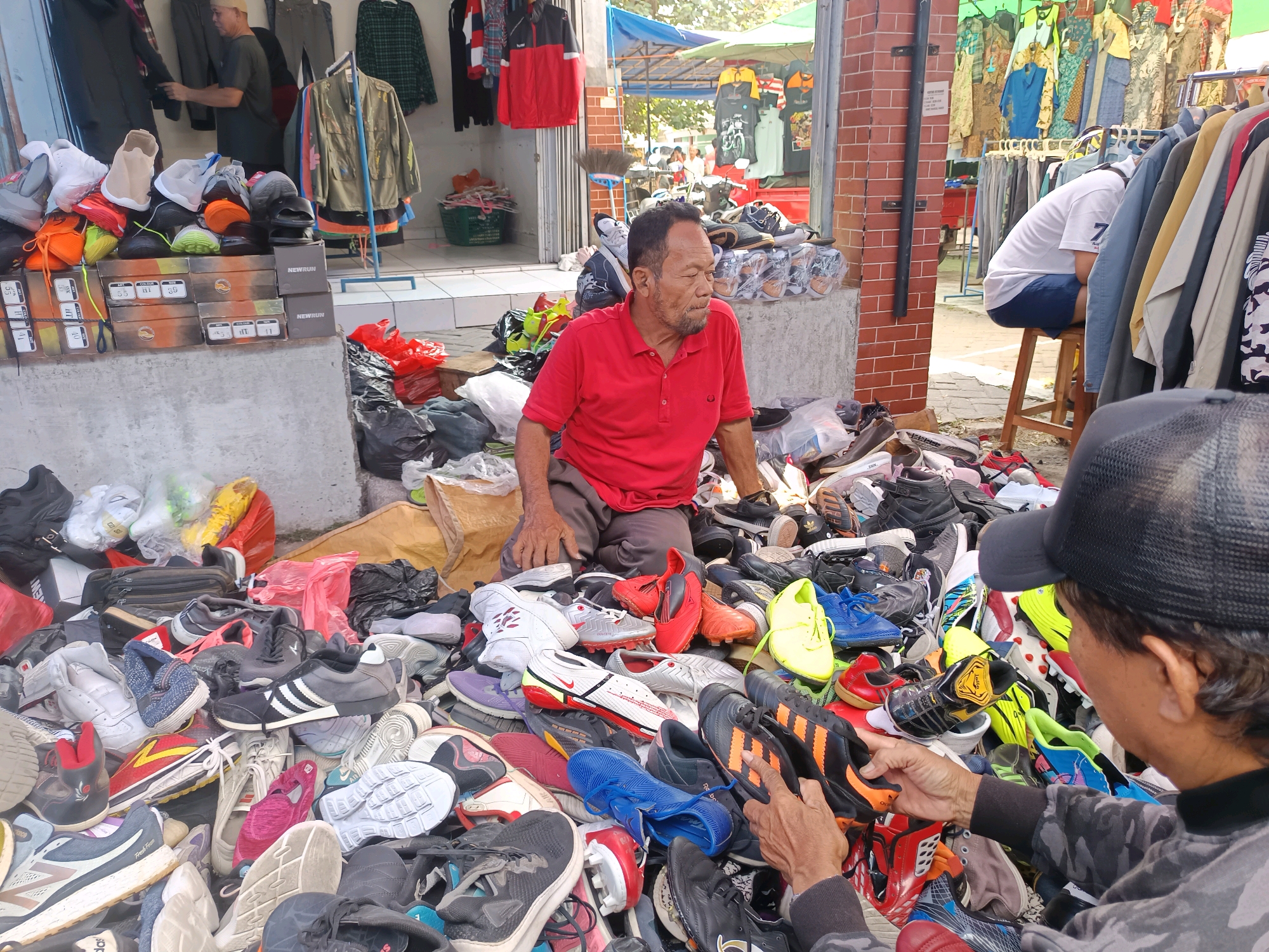 In Geneng, Indonesia, two men are seen at an outdoor market stall. The man in the foreground, wearing a red polo shirt, is seated among a large pile of shoes. He has dark hair and a beard, and appears to be a vendor. Behind him, a person wearing a white shirt and dark shorts is seated on a stool, partially obscured by hanging clothes. The stall is filled with various types of footwear, including sneakers and athletic shoes, displayed both on the ground and in boxes. Some of the shoe boxes have text that appears to be brand names or product information, such as "NEW RUN" and what looks like shoe sizes and colors. The backdrop reveals racks of clothing, suggesting the stall sells both shoes and apparel. The scene takes place during the day, with natural sunlight indicating it's likely morning or afternoon. The weather appears to be fair. The overall atmosphere suggests a bustling market environment.