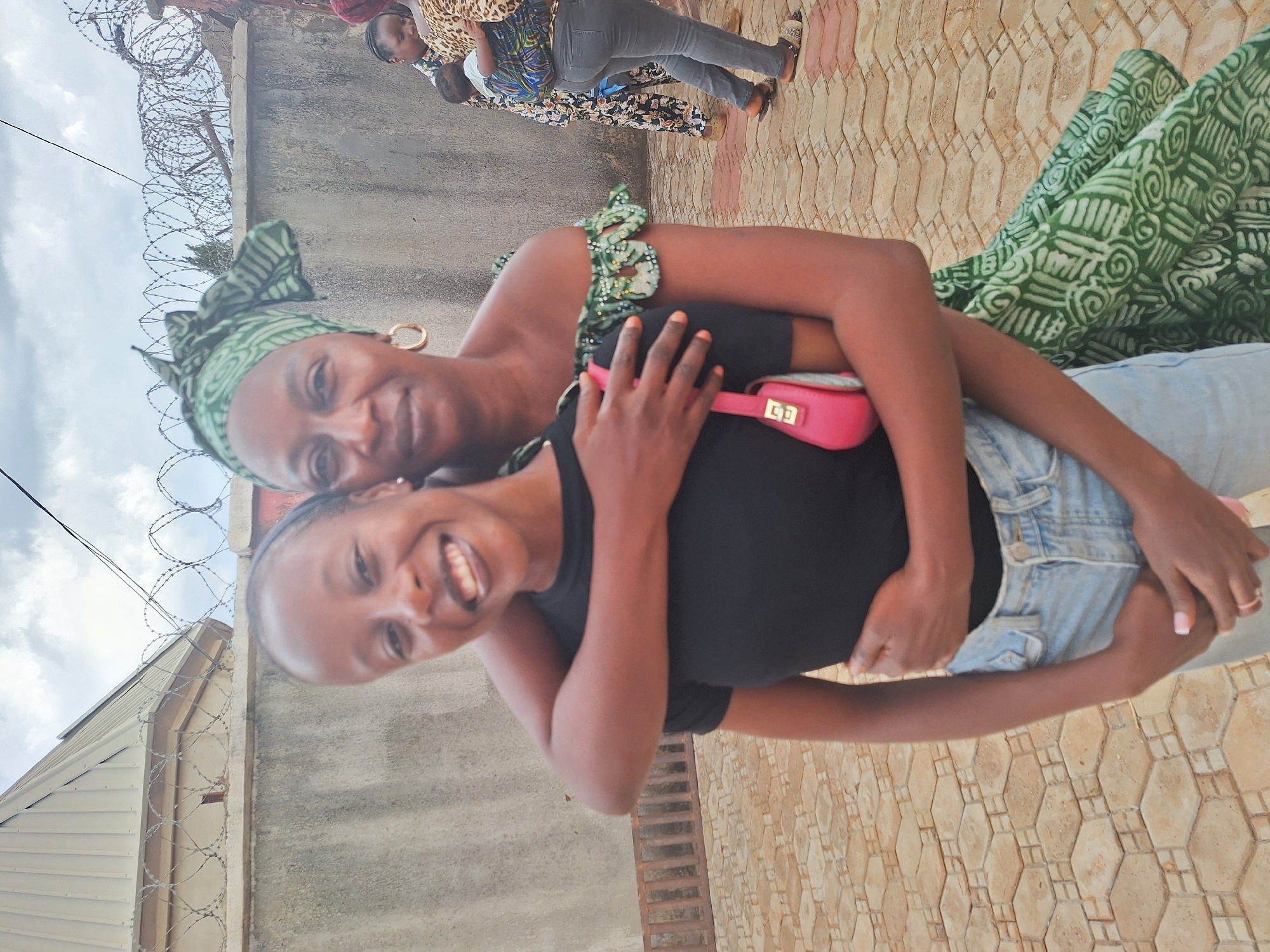 Two dark-skinned women are closely embracing outdoors. The younger woman wears a black top and denim jacket and smiles broadly. The older woman is behind her, with a green patterned headscarf and a green dress. The background shows a wall topped with barbed wire, the side of a building, and a textured brick-like ground surface. Other people are visible further in the background. The setting appears to be in Rido, Nigeria.