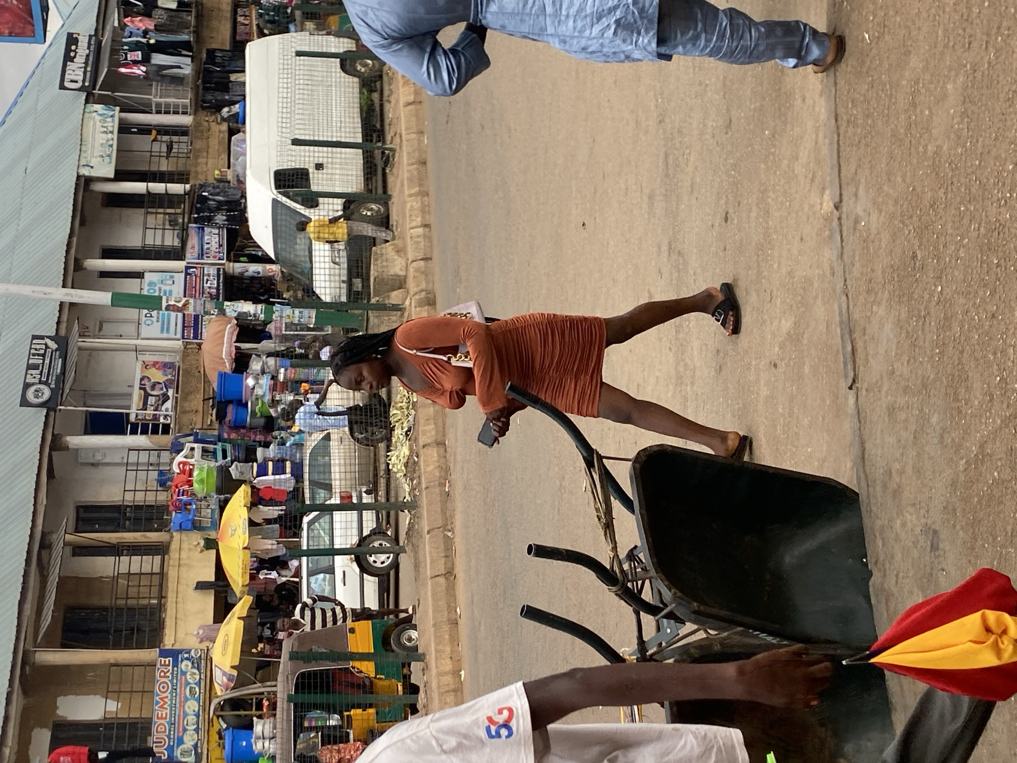 This image captures a bustling street scene in Bukuru, Nigeria, during daylight hours. The perspective is from slightly above, looking down a commercial thoroughfare.
In the immediate foreground, the arms and torso of a person wearing a white t-shirt with a blue and red "5G" logo on the shoulder are visible. This individual is pushing a dark-colored wheelbarrow, which has a red and yellow cloth draped over its handle.
The midground features a woman walking along the road's edge. She has dark braided hair, is dressed in an orange, ruched short dress, and carries a white shoulder bag. She is looking down at a smartphone held in her hand. Behind her, a man stands in a light blue traditional tunic and trousers.
The background reveals a lively market or commercial district. Buildings with corrugated metal roofs line the street, featuring multiple levels and barred windows. Various shops and stalls display merchandise, including clothes hanging on racks and stacks of colorful plastic containers. Several bright yellow market umbrellas are open, likely providing shade for vendors. A white van with a caged rear section is parked, and a yellow tricycle or "Keke Napep" is also visible further down the street. The road surface appears to be a mix of dirt and pavement.
Visible text includes "CBL Original" on a white sign attached to a building. Another prominent blue sign advertises "JUDEMORE GLOBAL INVESTMENT LIMITED," along with other text. A partially obscured sign reads "DEN OF CH..." and the "5G" logo is clear on the foreground person's shirt. The scene conveys everyday commerce and movement in an active urban environment.