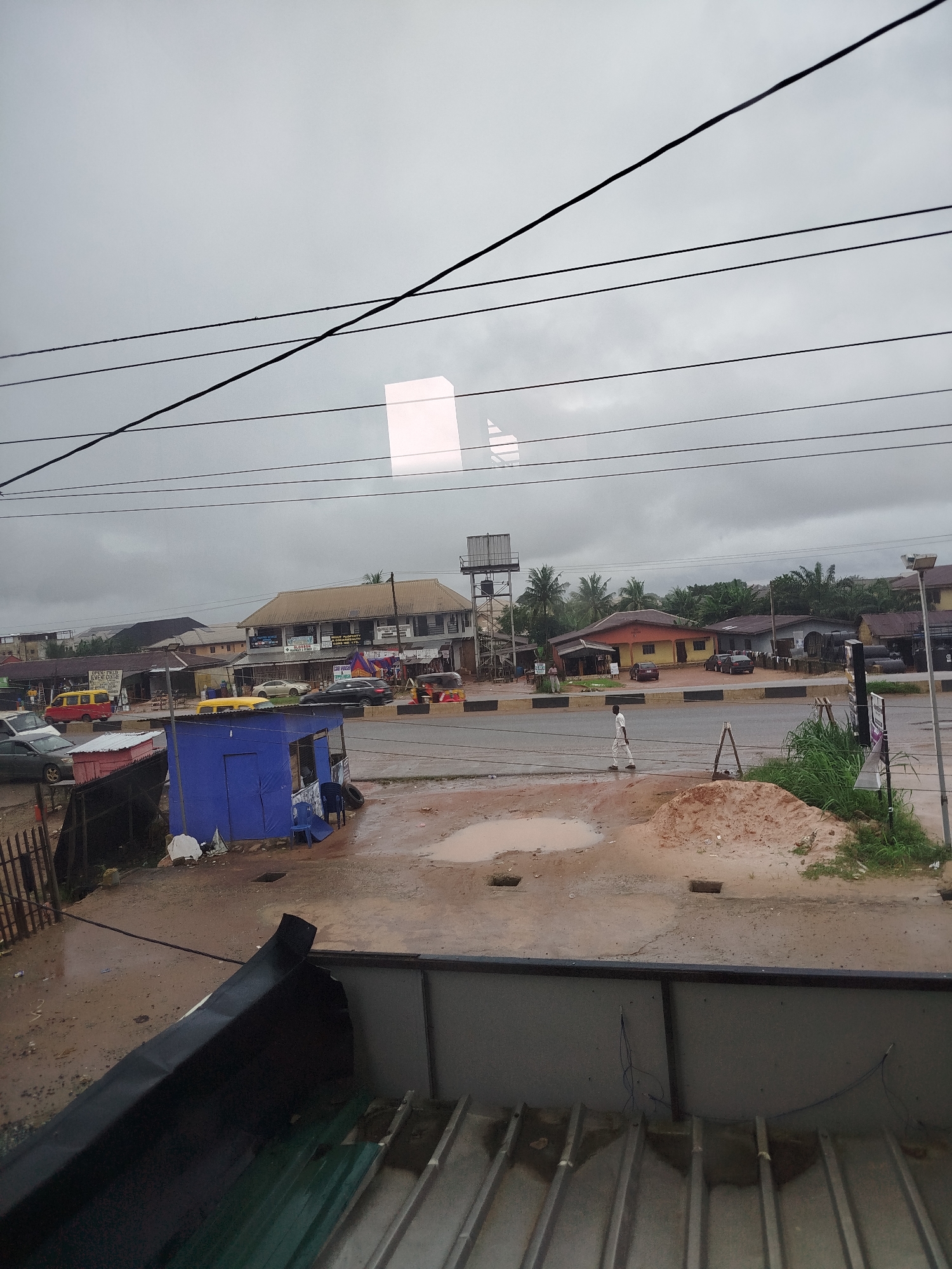 This image depicts a street scene in Uselu, Nigeria, under a cloudy sky. In the foreground, there's a portion of a corrugated metal roof. The middle ground shows a muddy road with several puddles, a pile of reddish-brown dirt, and a small blue makeshift structure with some debris around it. A lone individual wearing a white shirt and dark pants is walking on the road. Beyond the road, there are several buildings, including a long, two-story structure with shops on the ground floor. A yellow minivan and other vehicles are visible on the road and parked in front of the buildings. Telephone wires are strung across the upper portion of the image. The overall environment appears to be a commercial or residential area, possibly experiencing rainy weather given the wet road and puddles.