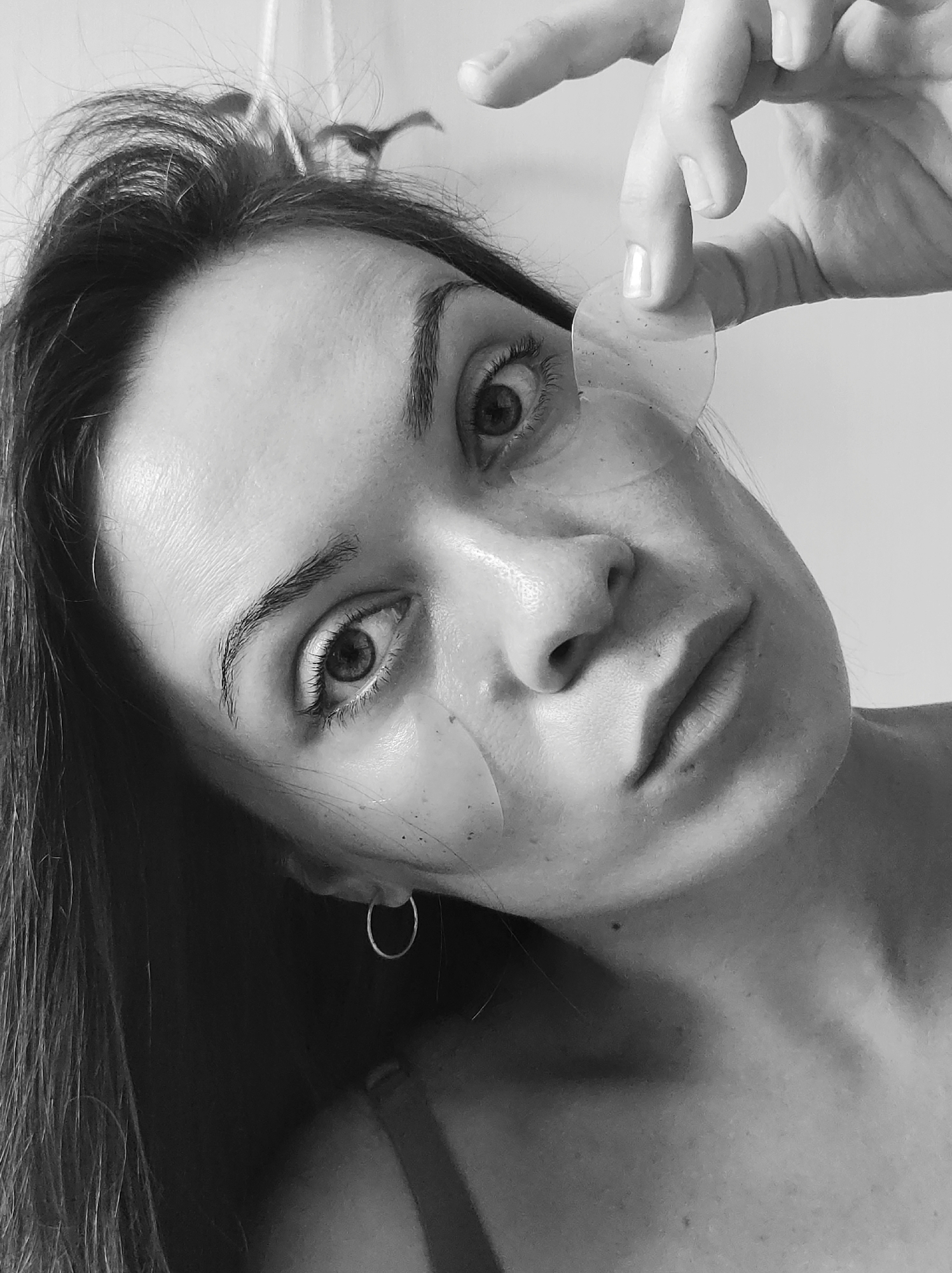 A black and white close-up photo shows a woman's face at an angle. She is holding a round object, possibly a skin patch, near her right eye. Another similar patch is already applied under her left eye. The woman has dark hair and eyes, with an earring visible in her left ear. The background is plain and light-colored, suggesting an indoor setting. There is no indication of the city of Chaikovsky, Russia in the picture.