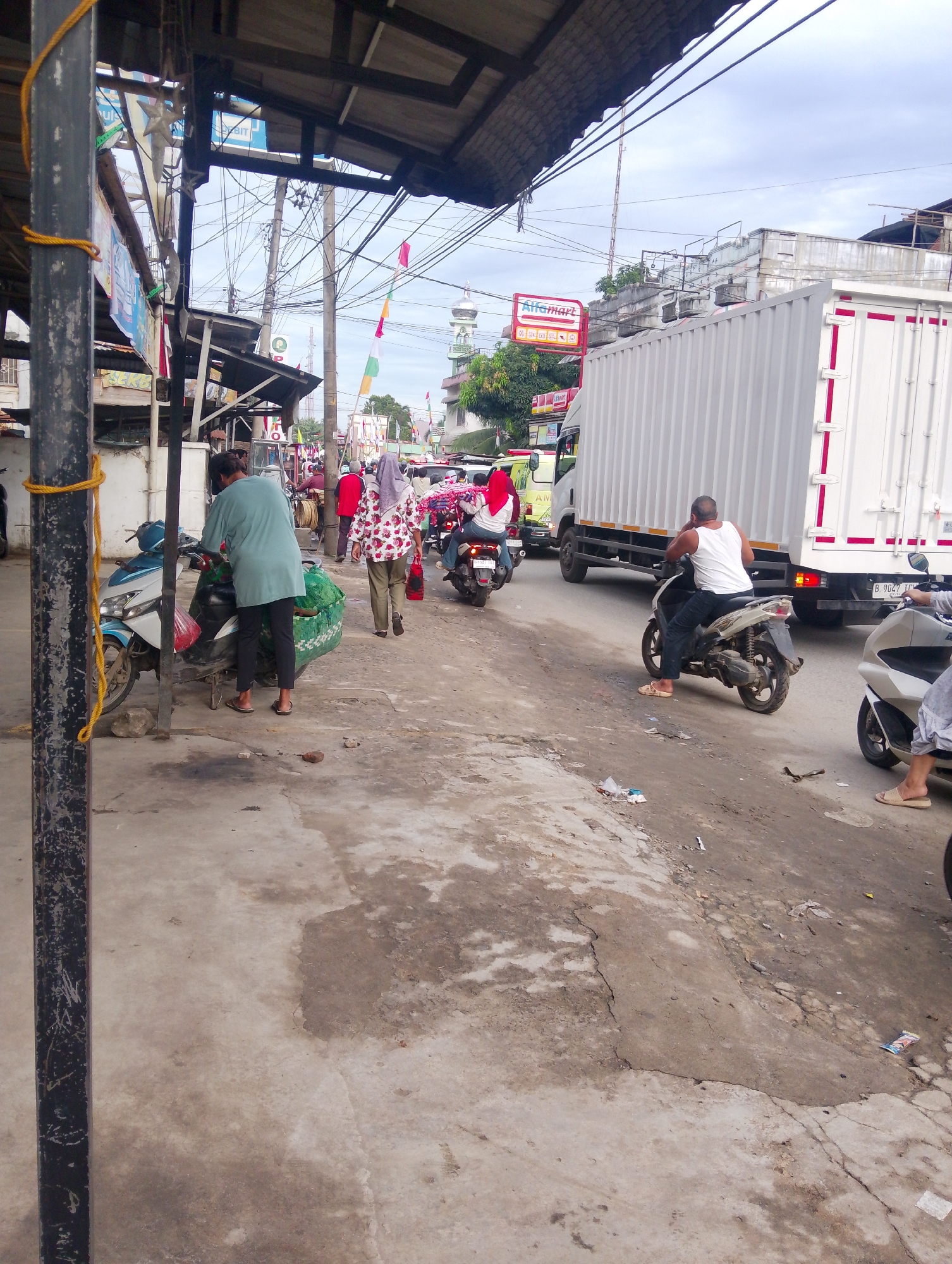 The image captures a street scene in Serbelawan, Indonesia, characterized by moderate traffic and pedestrian activity. A large white box truck is a prominent feature, positioned on the right side of the frame, its rear visible. To its left, a man wearing a white tank top and dark pants is seated on a beige scooter, facing away from the camera. In the foreground, a person in a light teal shirt and dark pants is attending to a blue and white scooter. This scooter is equipped with a large green woven bag containing what appears to be produce.
Further back, on the left side of the street, a woman in a patterned shirt with red and white floral motifs, wearing khaki pants and a red bag, walks away from the camera. Another individual in a red shirt is also visible in this area. Several other scooters and people are scattered throughout the scene, indicating a busy, possibly market, environment.
The environment consists of a paved road with some debris and patches of dirt. Overhead, a canopy structure extends from buildings on the left, supported by posts. Numerous electrical wires crisscross the sky, a common sight in urban areas. In the distance, a mosque with a silver dome is visible, along with other buildings and trees. The sky is overcast, suggesting cloudy weather. The presence of small flags and banners indicates a local celebration or event. The overall impression is one of a typical day in a bustling Indonesian town.