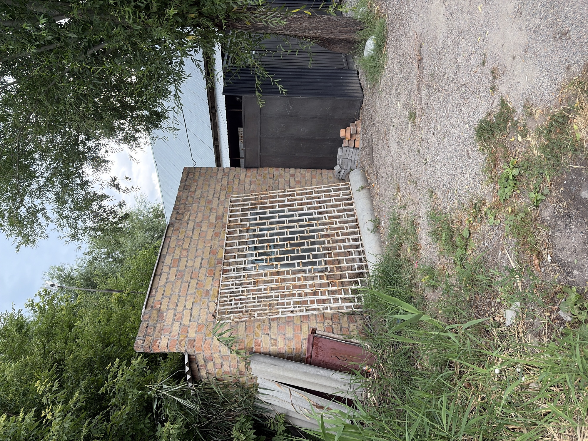 In the city of Kant, Kyrgyzstan, a close-up shot captures a scene near a brick building with a corrugated metal roof. In the foreground, tall green grass and some dry vegetation grow on a gravelly ground. To the left, several flat, gray construction panels lean against the brick building. Adjacent to them is a dark red rectangular object.
Dominating the center of the frame is a large, white, decorative metal grate, showing signs of rust, leaning against the brick wall. Behind the grate, a window with blue panes is visible in the brick structure.
To the right of the brick building, a tall, dark, ribbed metal gate stands closed. In front of this gate, a small stack of bricks is piled on the gravel. A mature tree with a thick trunk and green foliage grows to the right of the gate. The background reveals the sloped, light-colored metal roof of another building extending beyond the brick structure. The sky above is partly cloudy, with a light pole topped with a white object visible amongst the trees in the upper left portion of the image.