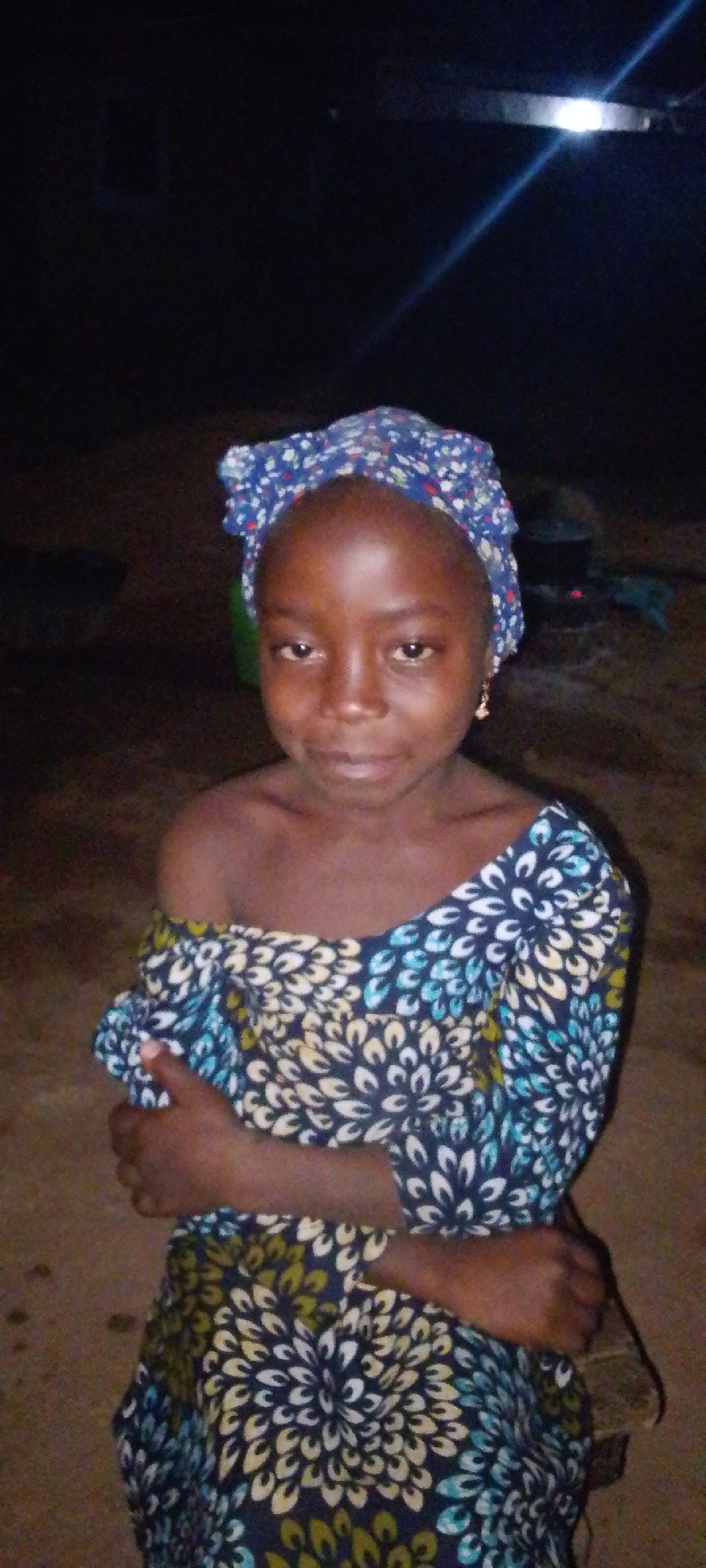 A young African girl stands with arms crossed in Jos, Nigeria, likely at night. She wears a floral print head wrap and matching off-the-shoulder dress. The background is dark, with a light source possibly illuminating the area above her. Two dark objects, perhaps cooking pots, are faintly visible in the background. The ground appears to be dirt.