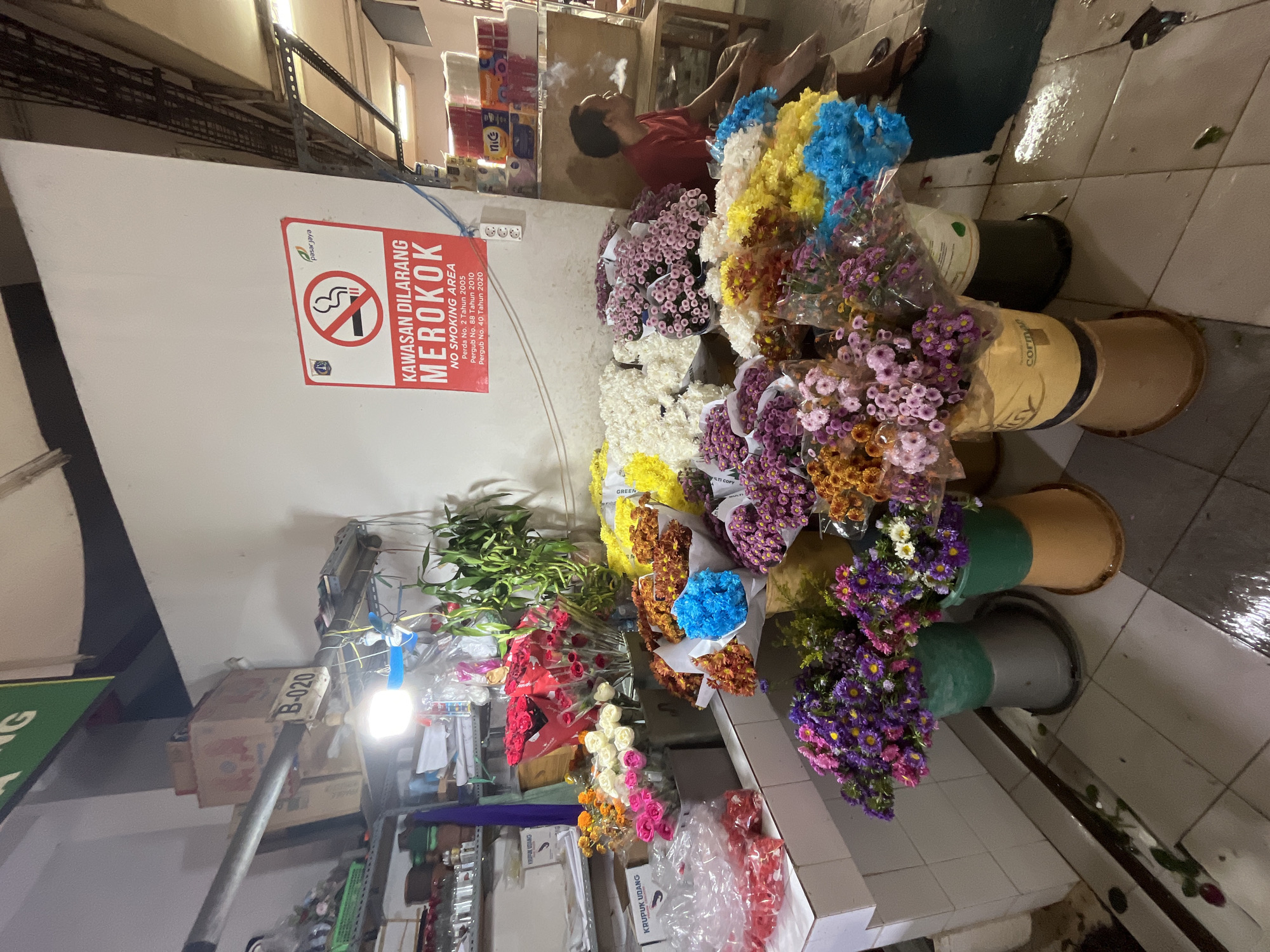 A man is smoking a cigarette in a flower market. He is wearing a red t-shirt and is seated on a wooden stool. In front of him are several buckets of colorful flowers, including red roses, white roses, pink roses, purple asters, yellow chrysanthemums, and blue asters. The man is smoking a cigarette, and there is a "No Smoking Area" sign on the wall behind him. The floor is tiled and appears wet in some areas. Shelves are visible in the background, with boxes and other items. The overall scene suggests a vibrant but somewhat chaotic market environment.