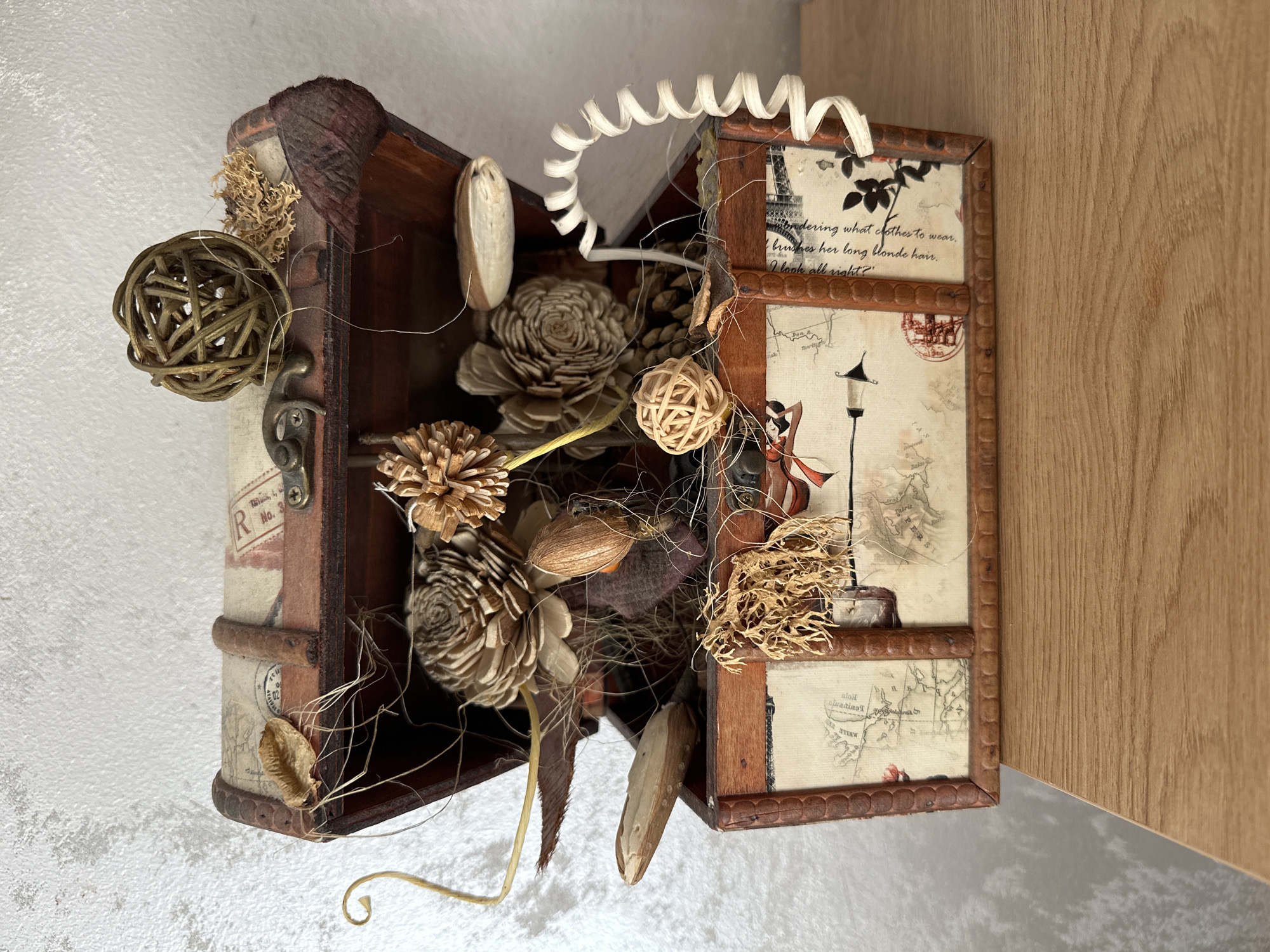 A decorative wooden chest, resembling a treasure chest, sits on a wooden surface against a textured grey wall. The chest is open, revealing an assortment of dried natural elements and decorative items. These include what appear to be dried flowers, pinecones, seed pods, and small woven balls. Some of these items are arranged within the chest, while others are attached to the exterior or lid. The chest itself has a rustic aesthetic, with decorative paper applied to its front and lid, featuring map-like designs and script. A coiled white decorative element extends from the right side of the chest. The lighting suggests an indoor setting. No people are present in the image. The image was taken in Glavatartsi, Bulgaria.