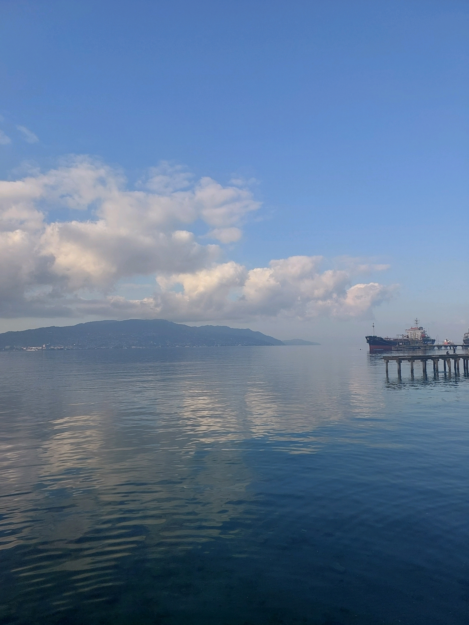 The image depicts a serene seascape with a calm body of water reflecting the sky and clouds. In the distance, a mountainous landmass, likely a part of Ambon Island in Indonesia, is visible. On the right side of the frame, a pier extends into the water, with a large cargo ship anchored nearby. The sky is a clear blue, with scattered fluffy white clouds. The overall impression is one of tranquility and natural beauty.