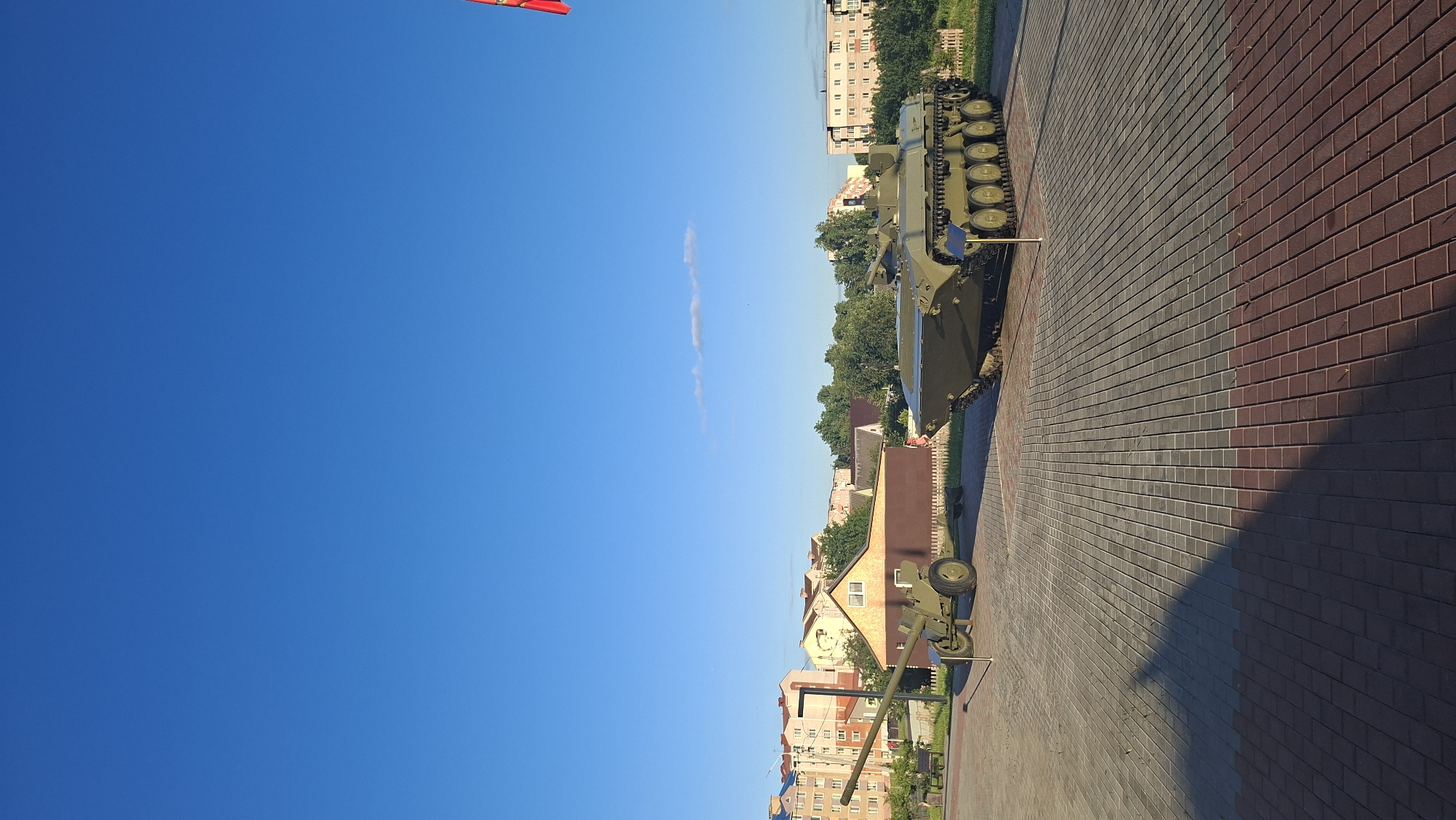 In the city of Smalyavichy, Belarus, a sunlit outdoor display features two military vehicles on a paved plaza. To the left, a green artillery piece rests on wheels. To the right, a green amphibious tracked combat vehicle is parked. Behind the vehicles, a low-rise building with a brown roof and beige walls stands, with residential buildings and lush green trees forming the backdrop under a clear blue sky. A red and yellow flag, partially visible at the top right corner, flutters in the breeze. The plaza is paved with gray and reddish-brown bricks, creating a distinct pattern. Shadows of the vehicles and surrounding elements stretch across the pavement, indicating it is daytime.