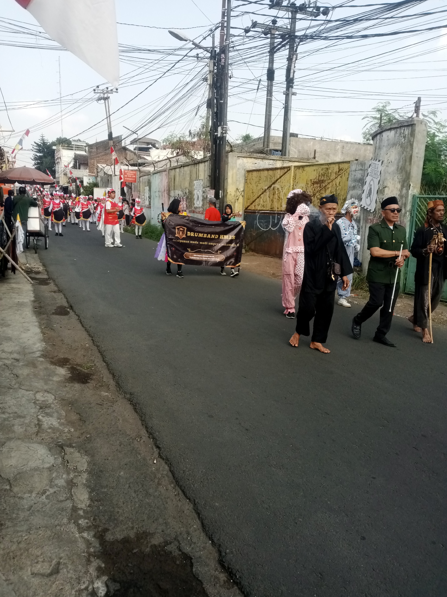 A group of people are participating in a parade on a street in Tasikmalaya, Indonesia. The parade features a drum band, some of whom are wearing red and white uniforms and carrying instruments. Several individuals are walking in front of the drum band, including two people in elaborate costumes, one with a pink polka-dot outfit and a large afro wig, and another with white face paint and a ruffled costume. A man in a black outfit is smoking a cigarette, and another man in a green uniform is holding a white stick. The street is paved and has power lines overhead. The buildings in the background appear to be commercial or residential structures. The event seems to be taking place outdoors during the day.