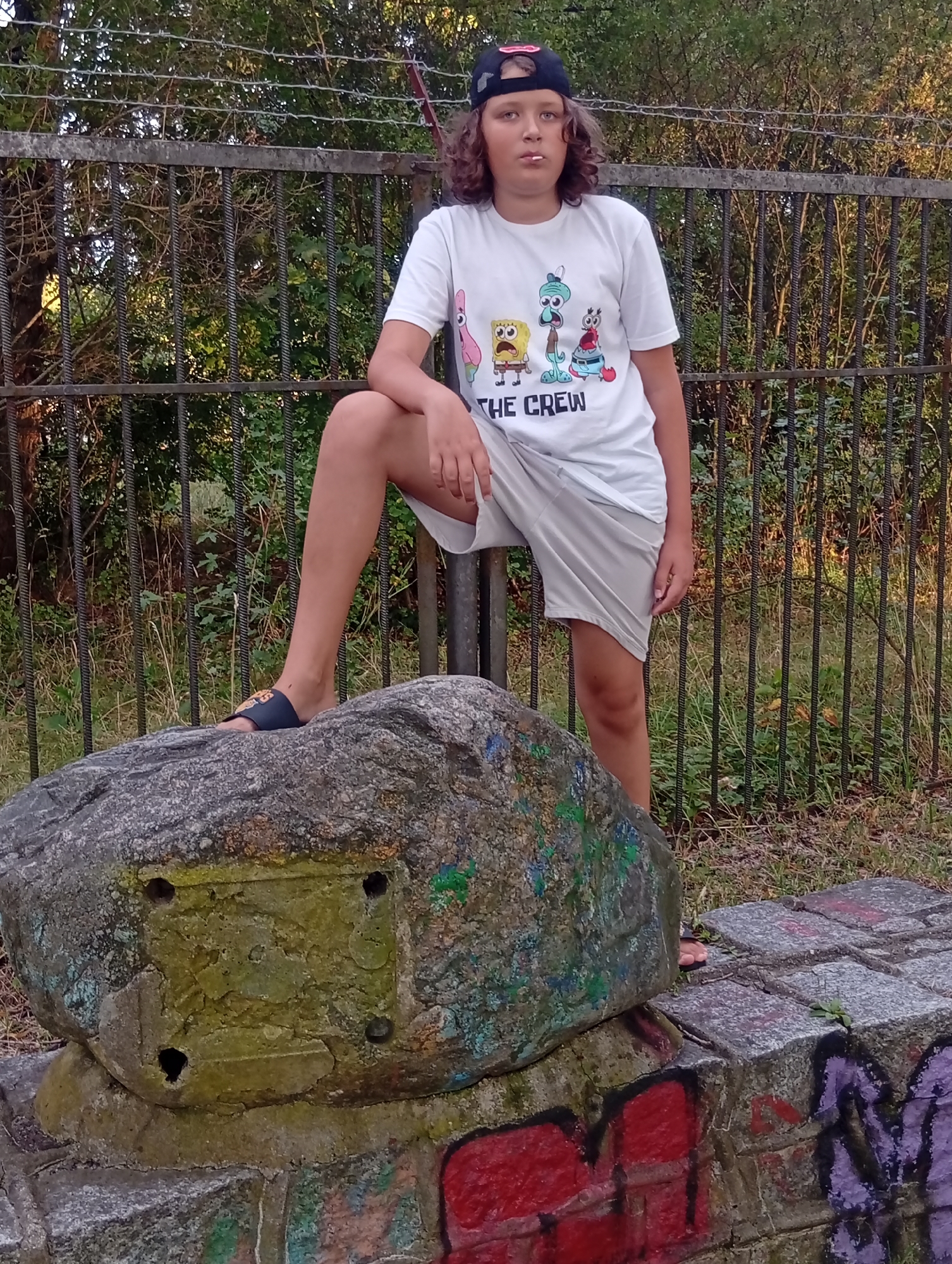 A young person with curly brown hair and a black baseball cap is standing with one leg on a large boulder. They are wearing a white t-shirt with cartoon characters and the words "THE CREW" printed on it, and light-colored shorts. The boulder is positioned on top of a graffiti-covered stone structure. Behind the person is a metal fence with barbed wire at the top, and trees and foliage visible beyond the fence. The ground around the boulder and structure is covered in grass and dirt. The scene appears to be outdoors in a public space, possibly a park or recreational area. The lighting suggests it is daytime. The presence of graffiti and the weathered appearance of the fence and boulder indicate the location may be somewhat neglected. No specific landmarks or indications of Wrocław, Poland, are visible in the image.
