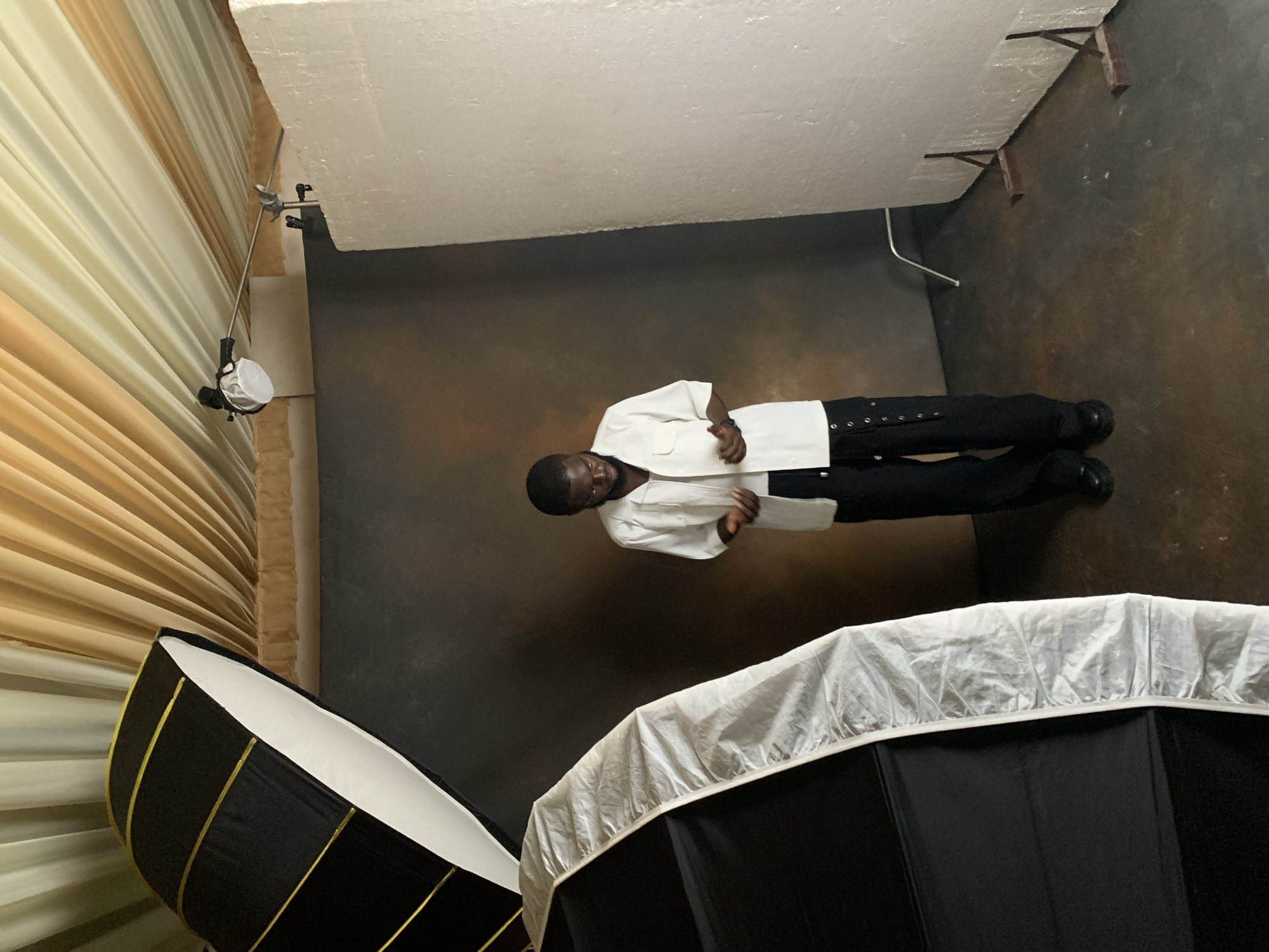 A Black man stands in a photo studio in Katon Rikkos, Nigeria, looking down at his hands. He is wearing a white short-sleeved button-up shirt and black pants with cargo pockets. He has dark hair and is wearing glasses. The studio features a dark backdrop, a large softbox to the left, a large white styrofoam board to the right, and draped yellow and cream fabric on the ceiling. A small light fixture is mounted on the ceiling. The floor is concrete.