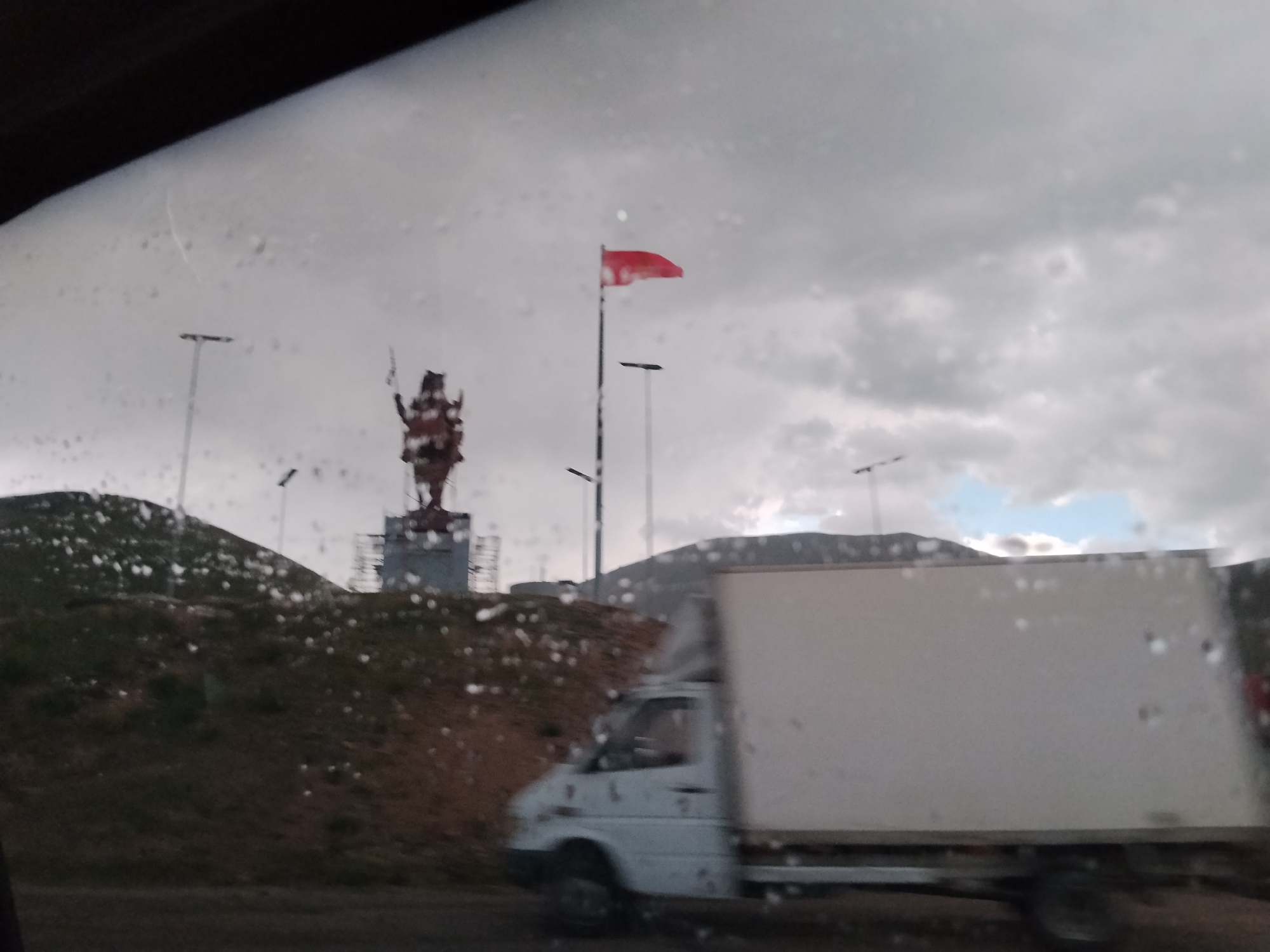 The image depicts a cloudy day scene viewed from a vehicle, likely a car due to the perspective and the presence of water droplets on the glass. In the background, a statue of a figure with multiple arms is visible on a hill, with scaffolding around its base, suggesting ongoing construction or maintenance. A red flag is mounted on a pole to the right of the statue. Further in the background, a hazy mountain range is discernible. In the foreground, a white truck is in motion, its side largely obscuring the view of the road and surrounding terrain. The terrain itself appears to be hilly with sparse vegetation. The scene is set in a location that is identified as Unknown, Kyrgyzstan.