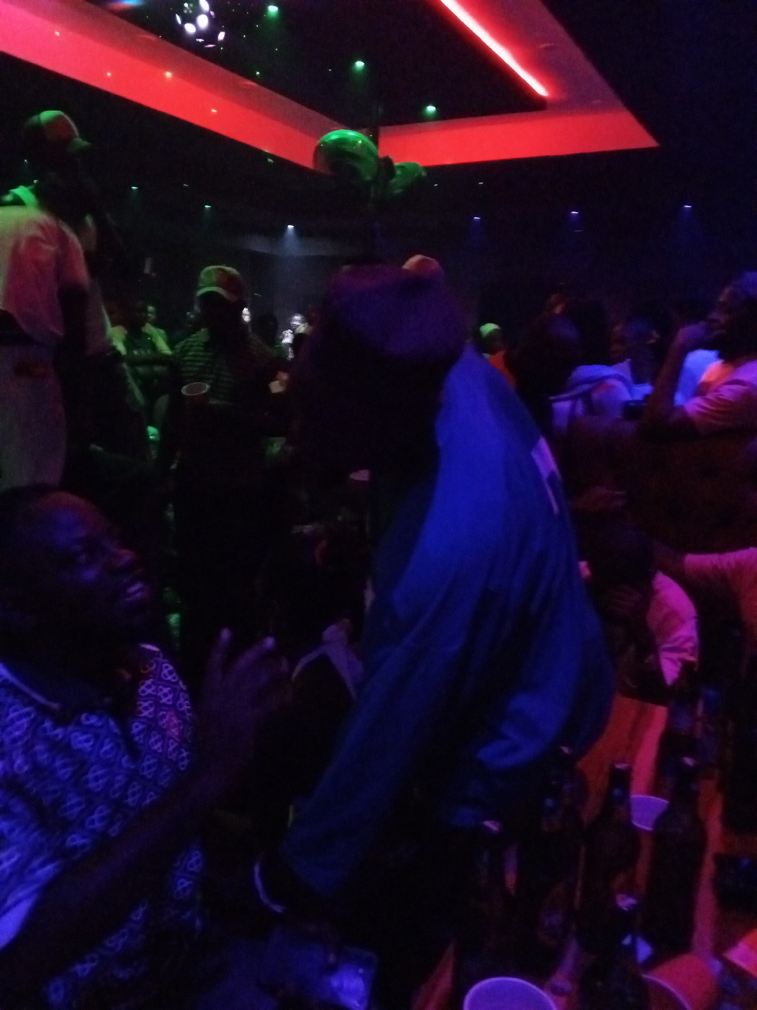 A dimly lit indoor scene, possibly a nightclub or bar in Lokoja, Nigeria, shows several people engaged in social activity. Individuals are seen standing and sitting, with some appearing to clap. There are bottles and plastic cups on a table. The environment is illuminated by colored lights, predominantly red and blue, creating a dynamic and lively atmosphere.