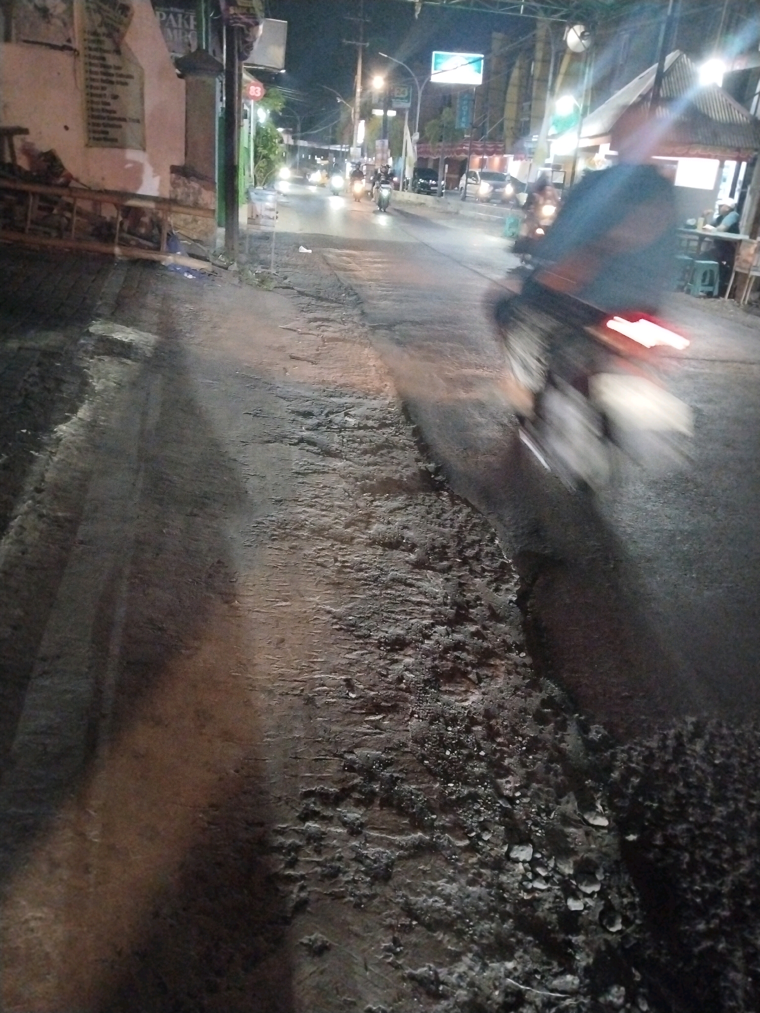 This is a nighttime image of a street in Pepelegi, Indonesia. The scene is dimly lit by streetlights and the headlights of vehicles. The foreground shows a rough, textured pavement with debris. A dark, blurry shadow of a person on a motorcycle is prominent on the right side of the image, with the motorcycle's taillight creating a red streak of light due to motion blur.
In the background, several vehicles, including motorcycles and cars, are visible on the road, their lights blurring due to the low light conditions and the camera's exposure. There are buildings and structures lining the street. One building on the left has some posters or signs on its wall. On the right, there appears to be a small open-air establishment with a roof, where a few people are seated at tables.
The overall atmosphere is that of a busy street at night, with traffic and activity. The motion blur suggests that the photo was taken with a slower shutter speed to capture the movement of the vehicles. The time of day is clearly night, and there are no strong indicators of specific weather conditions, but the wetness of the road in some areas could suggest recent rain. The visible text "PAKE" on a sign on the left side provides a minimal textual element to the scene.