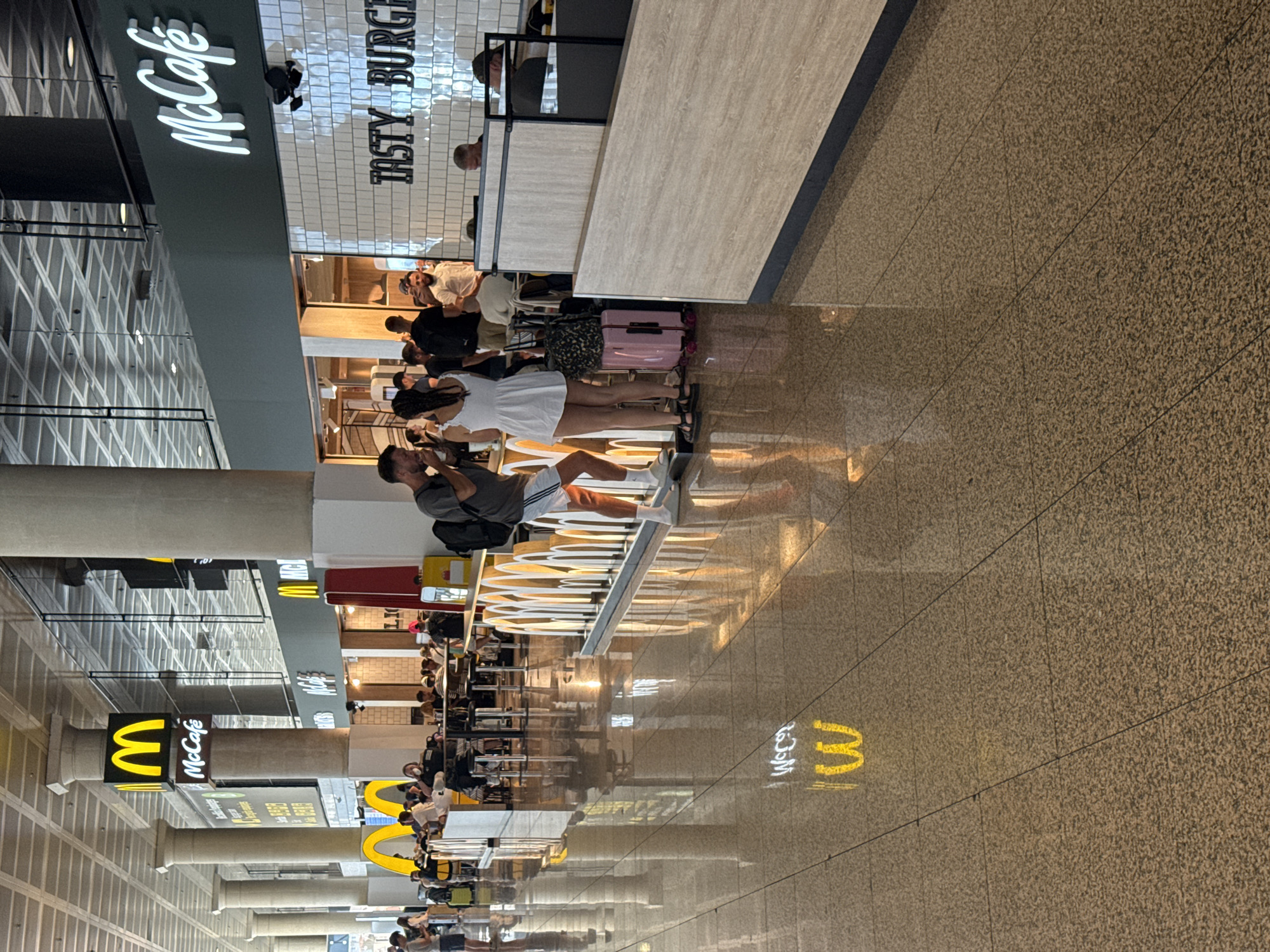 This image depicts a scene inside a McDonald's restaurant, likely located in an airport terminal at El Prat de Llobregat, Spain, given the presence of numerous travelers and the polished, spacious interior indicative of such a location.
In the foreground, a man wearing a grey t-shirt, white shorts, and white socks with grey slides is standing, facing to the right. He has a backpack over his right shoulder and appears to be holding something in his right hand, possibly food or a drink. To his left, a woman in a white dress with thin straps and black sandals is standing, facing towards the man. She has braided hair and is looking down, holding a white object in her left hand. Between them, a pink suitcase with black wheels is placed on the floor.
Behind them, a counter area is visible with several people queuing or sitting at tables. A sign with the "McCafé" logo is prominent above the counter. Further back, more seating areas and patrons are visible, along with other McDonald's branding. The floor is highly reflective, showing the illuminated McDonald's "M" logo from the ceiling as a bright yellow reflection.
The environment appears to be an indoor public space, likely a commercial area within a transportation hub. The lighting is bright, and the architecture features large pillars and a high ceiling with visible structural elements. The overall atmosphere is one of a busy commercial establishment catering to travelers.