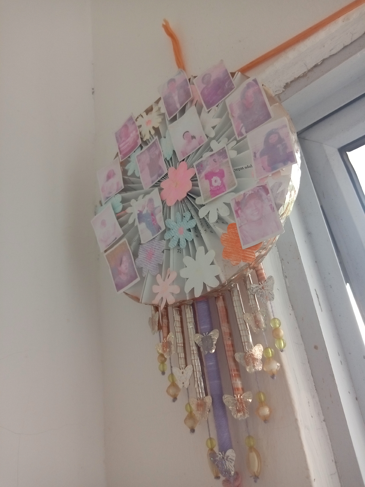 The image shows a DIY dreamcatcher hanging on a wall next to a window. The dreamcatcher is circular with a fan-like structure made of folded paper or fabric, adorned with small printed photographs of people, many of whom appear to be children. There are also decorative paper flowers in various colors, including pink, white, and light blue, attached to the dreamcatcher. Hanging from the bottom of the dreamcatcher are several strands made of beads and clear plastic butterfly charms, interspersed with what appear to be small rolled-up papers or tubes. The wall is plain and white, with a slight crack visible. The window frame is white and appears to be made of PVC. The light suggests it is daytime, with natural light streaming in from the window. The location is described as Batam, Indonesia, but there are no specific visual cues in the image that would confirm this geographical location; it is presented as a contextual detail. The overall scene suggests a personal, handcrafted item displayed in a domestic or casual setting.