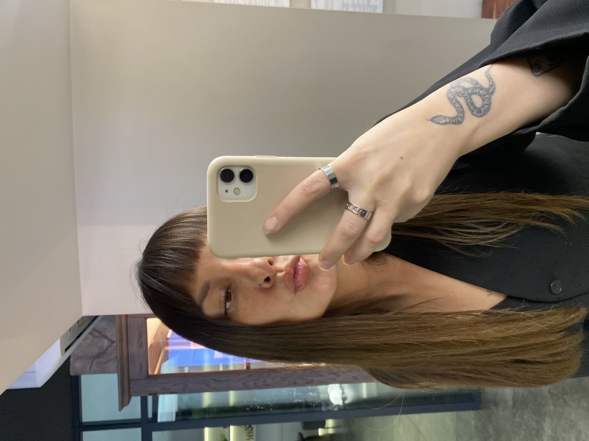 A selfie is taken by a woman with long, brown hair and bangs, wearing a black jacket over a dark top. She is holding a light-colored smartphone in her right hand, angled to capture her reflection. Her lips are pursed, and she is wearing a nose ring and two silver rings on her fingers. A snake tattoo is visible on her left forearm. The background is blurred, but appears to be an indoor setting with neutral-colored walls. There is a wooden cabinet with glass doors and lighting visible on the left side of the image, and a hint of a window with vertical blue and white stripes on the far left. No specific Votkinsk, Russia landmarks are identifiable in this image.