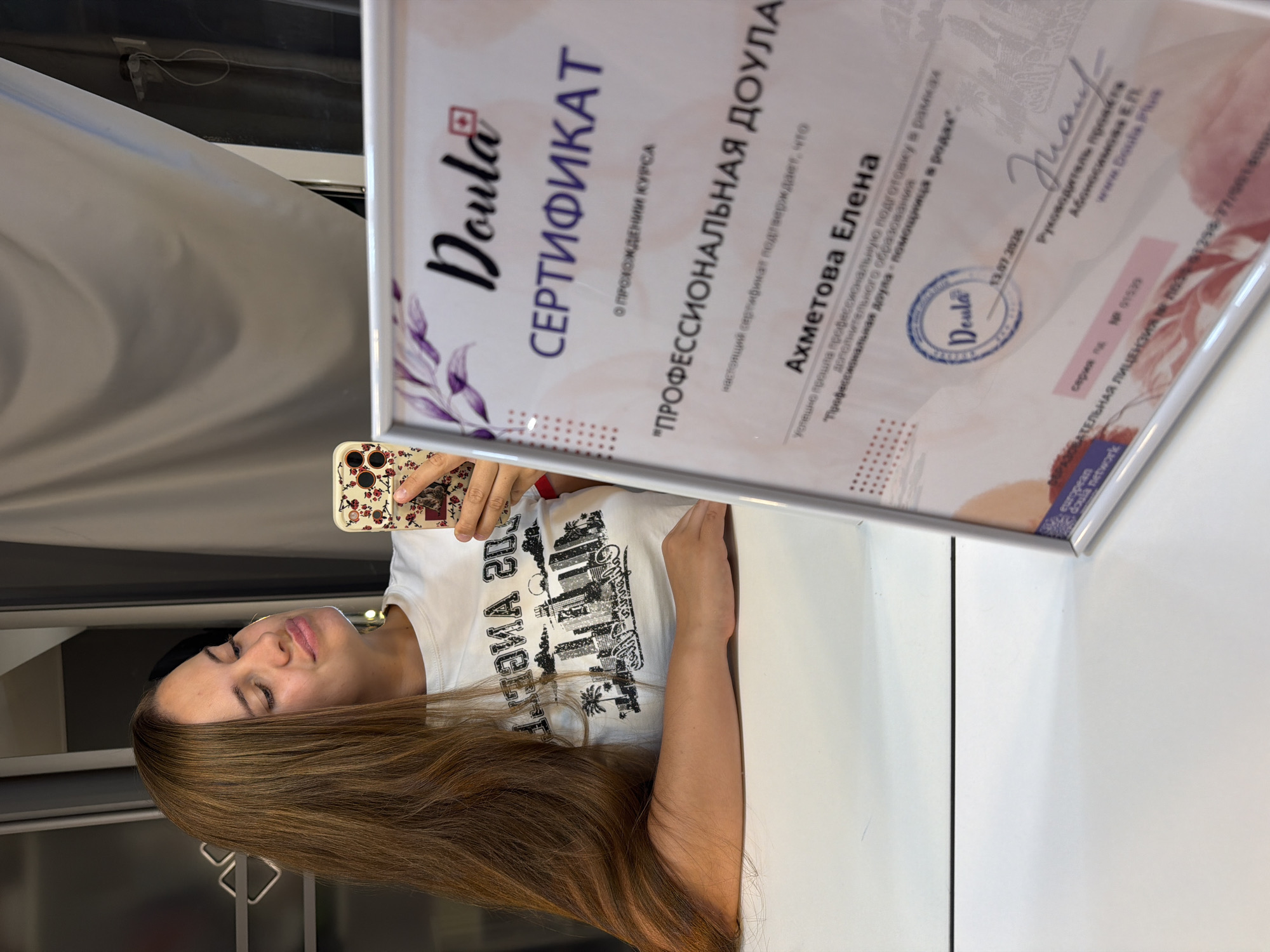 A woman with long, brown hair is taking a selfie in a room, possibly an office or studio. She is wearing a white t-shirt with a graphic print. In the foreground, a framed certificate is visible. The certificate appears to be a diploma or certification, with text in Russian and a seal. The text on the certificate indicates it is for a "Professional Doula" course. A stamp on the certificate reads "Doula" and a date, "13.07.2026". The background is blurry, but suggests an indoor environment with some curtains or drapes. The certificate has a decorative border with floral elements. The woman's posture and the selfie angle suggest she is posing to showcase the certificate. The presence of a professional certification suggests this is likely a business or professional setting related to doula services. There is no information in the image to indicate the location is Sochi, Russia.
