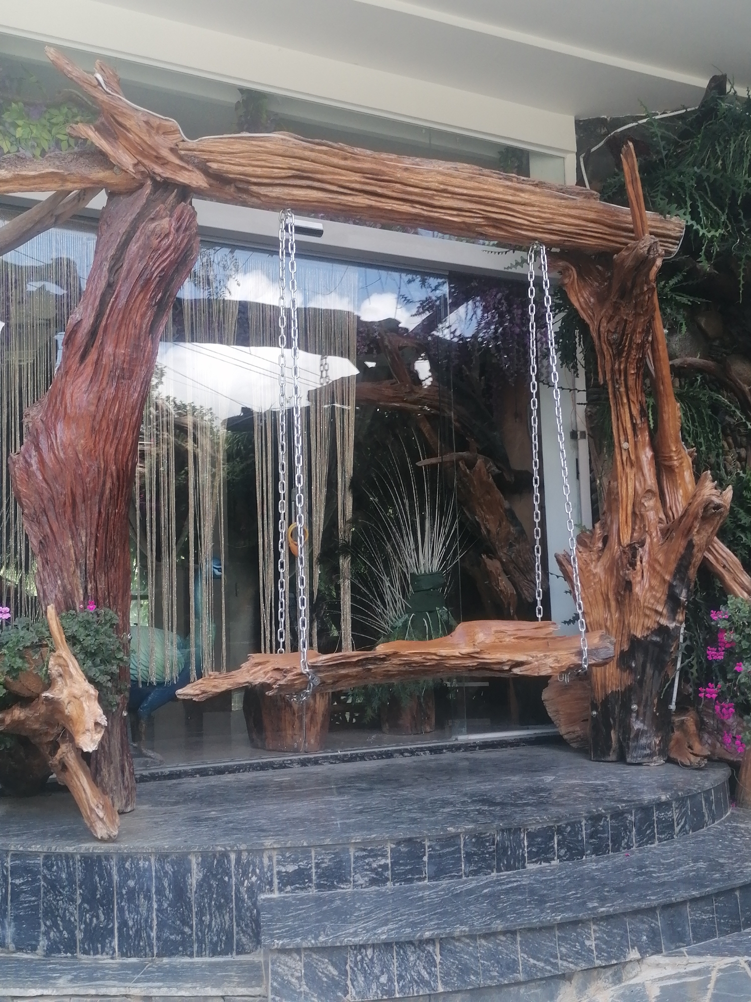 The image shows a unique swing set crafted from weathered tree branches, positioned outside a building with large glass windows. The swing features a rough-hewn wooden seat suspended by chains from a horizontal branch supported by two large, gnarled tree trunks. The backdrop through the glass reveals a dimly lit interior with vertical strands of decoration and some greenery. In the foreground, dark, textured stone steps lead up to the swing area, with small pink flowers blooming on the left and right sides of the swing. This rustic art installation, likely a decorative piece, is situated in Da Lat, Vietnam, a city known for its natural beauty and artistic expression, suggesting it could be part of a cafe, garden, or unique architectural space.
