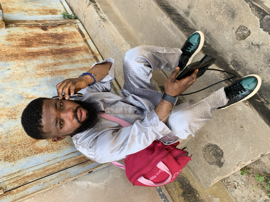 An adult male is seated on weathered concrete steps in Okene, Nigeria. He wears a light grey two-piece garment with a subtle dotted pattern, black and teal patent leather sneakers with white soles, a black smartwatch on his left wrist, and a blue beaded bracelet on his right wrist. A red "Hello Kitty" brand bag with a pink strap is slung over his left shoulder. He holds a black smartphone to his right ear with his right hand, indicating a phone conversation. His left hand rests on his left thigh, holding a black wallet or phone case with a strap. Behind him is a large, severely rusted metal surface, likely a gate or door, exhibiting extensive brown and orange oxidation. A small green plant grows from a crack in the concrete near the rusted surface on the right side of the frame.
