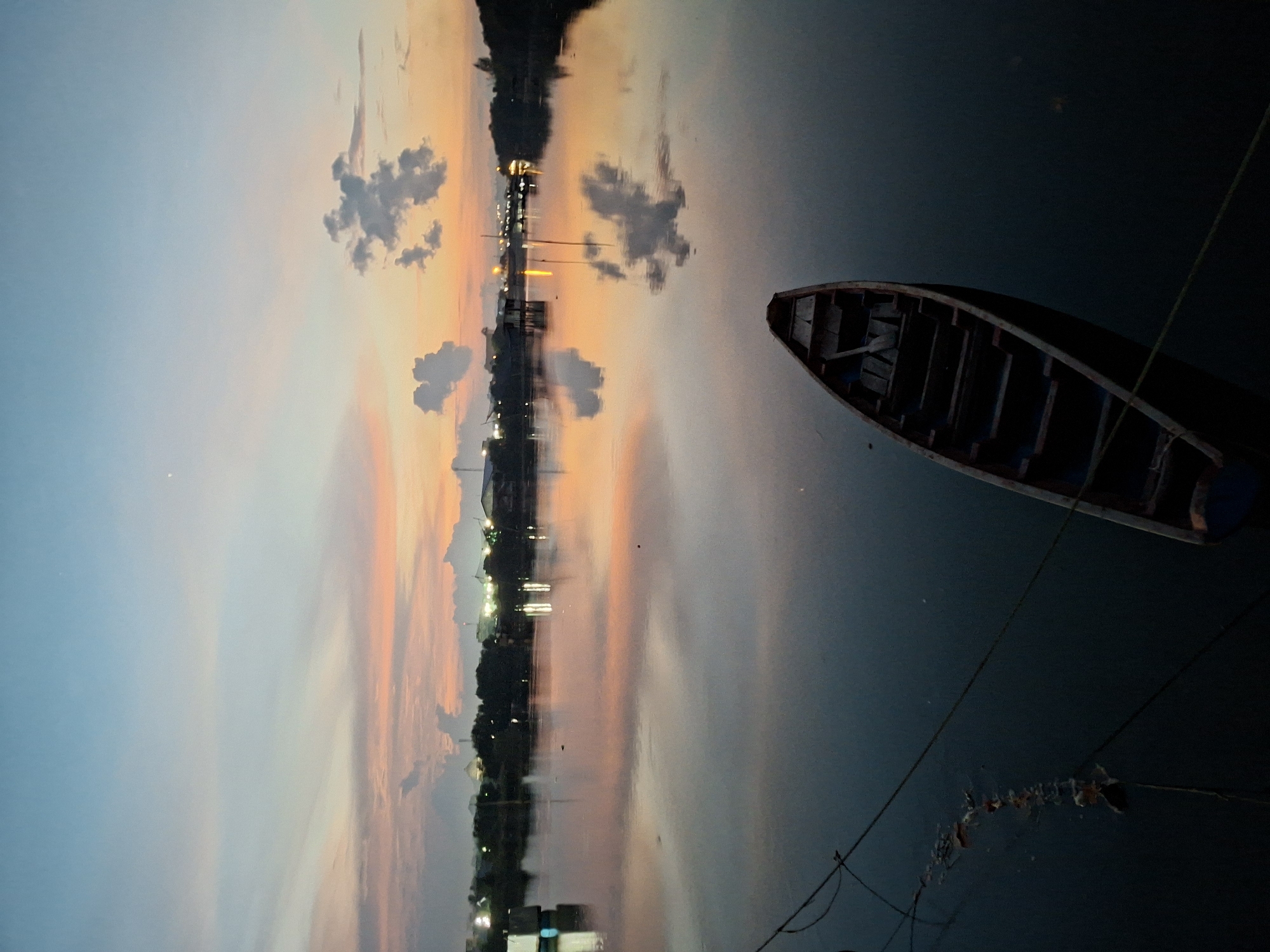 The image, presented with a 90-degree clockwise rotation, captures a tranquil waterscape at either dusk or dawn. In this rotated view, a dark, empty, traditional wooden boat is positioned on the right side of the frame, appearing to float vertically on the still water. Ropes, possibly mooring lines, extend from the boat towards the bottom right.
The central and upper sections of the rotated image showcase the near-perfect reflection on the water's surface. This reflection mirrors a sky displaying the soft blues, oranges, and pinks characteristic of sunset or sunrise, along with scattered dark clouds. In what would be the distant shoreline of a correctly oriented scene (visible in the lower-central part of the rotated image), a settlement or village is discernible. Its illuminated artificial lights cast bright, elongated reflections across the calm water, contributing to the peaceful atmosphere. The scene suggests a quiet moment by a body of water, such as a river or lake, consistent with environments found in Lhokseumawe, Indonesia. There are no people visible in the image, and no discernible text.