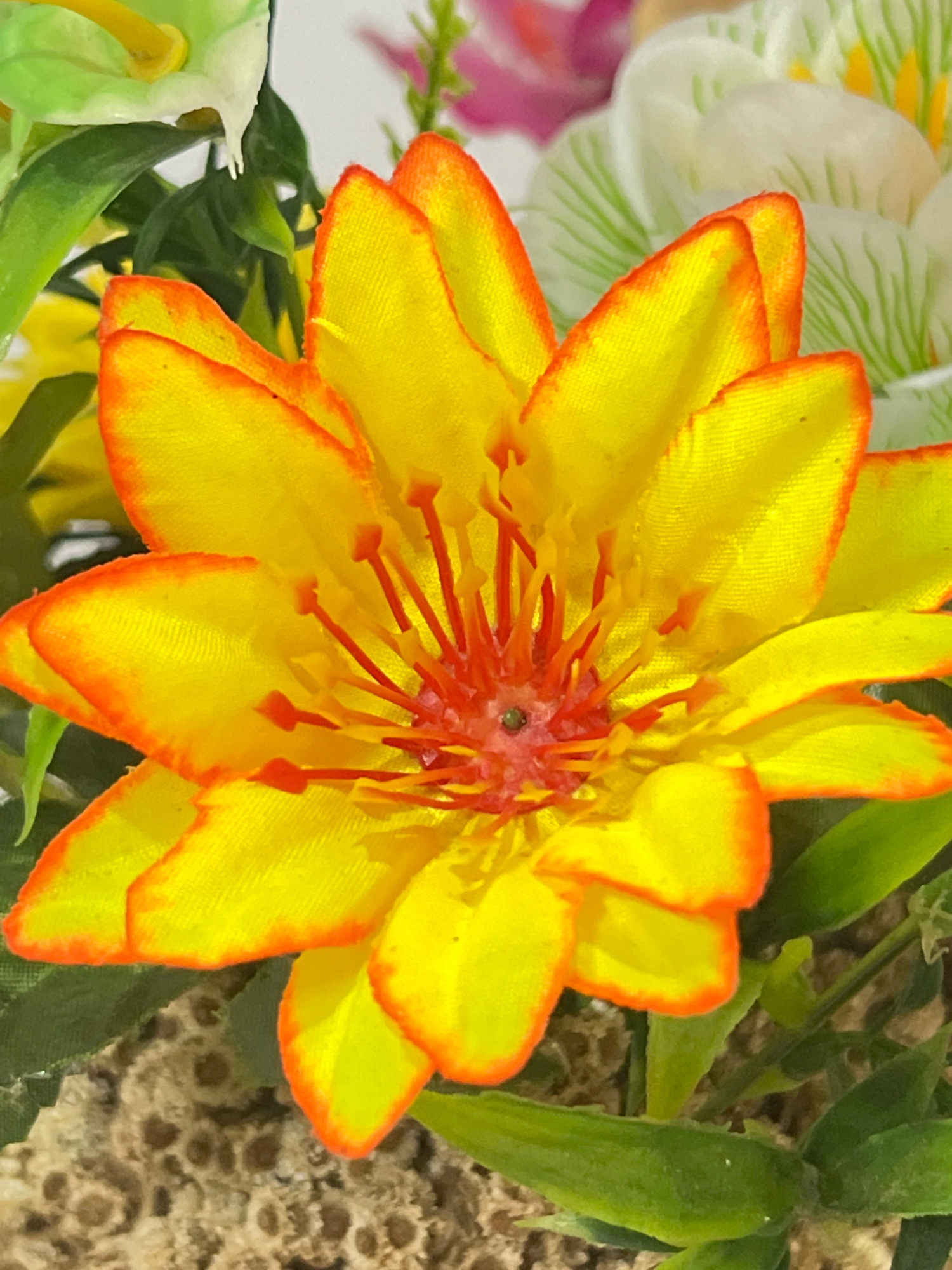 A close-up, eye-level shot captures a vibrant artificial lotus flower, primarily yellow with orange-red edges on its petals. The center of the flower displays numerous thin, red stamens tipped with small orange points, surrounding a dark, circular pistil. Behind the main flower, other artificial flowers are visible, including soft green and white blossoms with green veining, and hints of pink in the background. The texture of the petals appears fabric-like. The lower portion of the image shows green artificial leaves and a textured brown surface that resembles soil or a porous material. This image was taken on June 15, 2024, at 8:52 AM in Denpasar, Indonesia, at coordinates 8.6647° S, 115.2170° E.