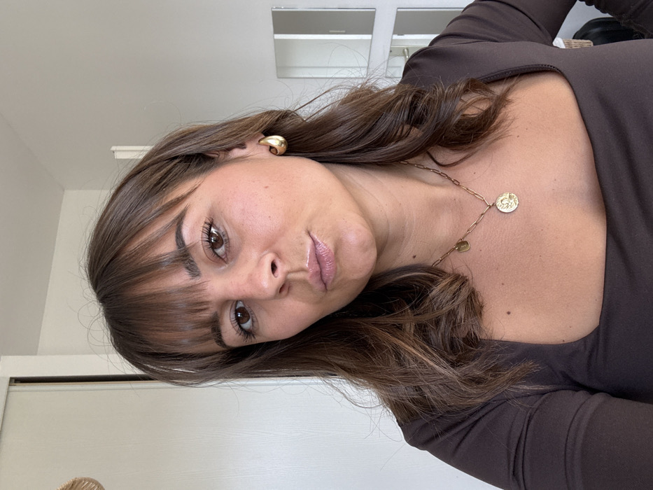 The image depicts an adult female posing for a close-up photograph indoors, located in Veauchette, France. The subject has long, wavy brown hair with bangs covering her forehead, brown eyes, and a tanned complexion. Her lips are slightly parted and puckered. She wears a dark brown long-sleeved top with a square neckline. A gold-colored hoop earring is visible on her right ear. Around her neck are two gold-colored necklaces: a paperclip chain with a prominent round medallion pendant featuring an embossed design, and a finer chain with a smaller oval pendant. The background consists of a light-colored wall with a vertical seam or door frame on the left. On the right, reflective panels with white framing are visible, suggesting a mirror or closet doors. A partially visible, light-colored woven circular object hangs on the upper left wall.