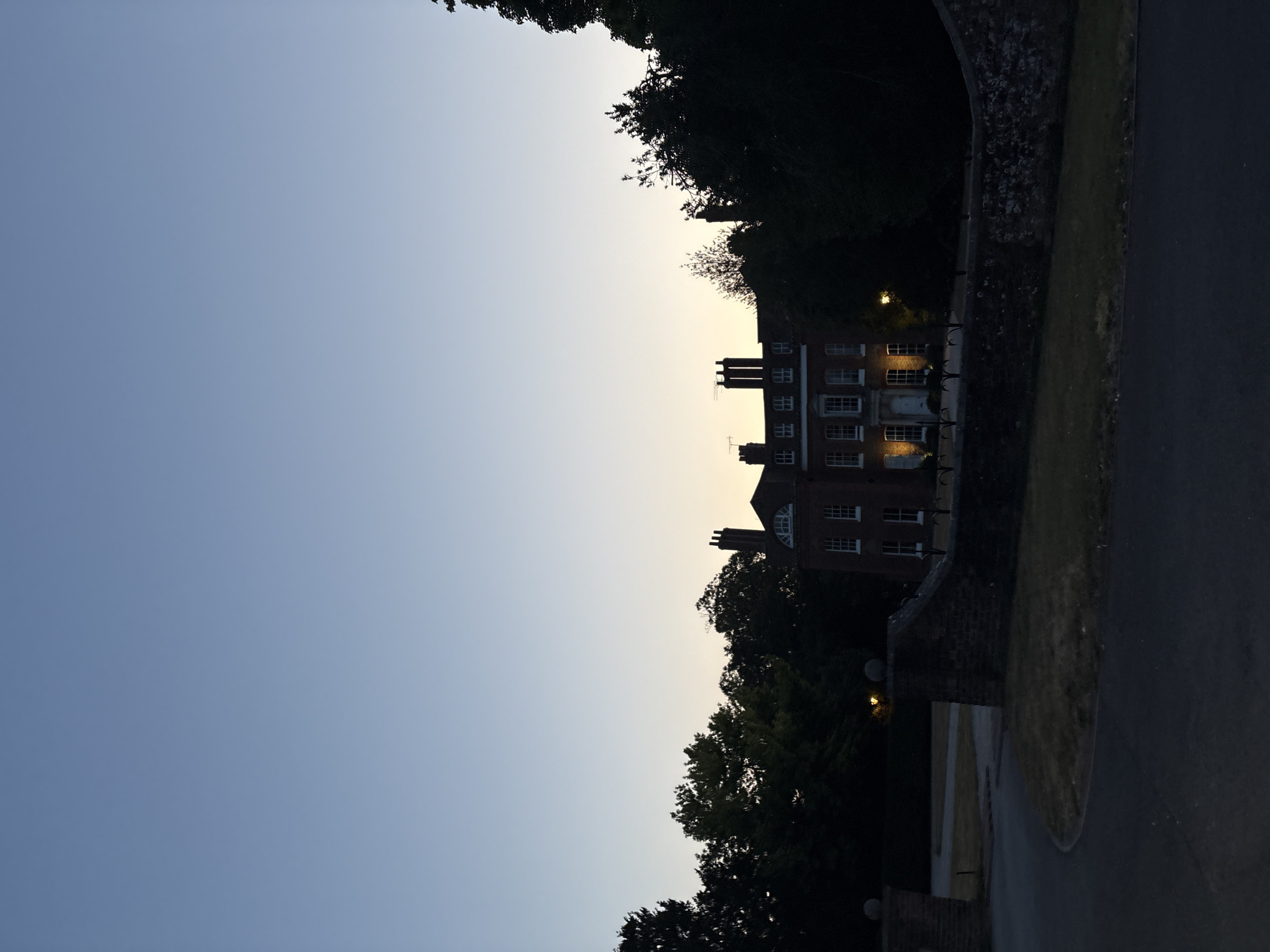 A stately brick house, with multiple chimneys and arched windows, is partially visible behind a stone wall and silhouetted trees. The sky above is a gradient of deep blue to pale yellow, suggesting either dawn or dusk. A paved driveway curves in the foreground. The house is lit by exterior lights. The scene appears to be located in a rural or suburban area, possibly on the outskirts of Chelmsford, United Kingdom. There are no people visible in the image.