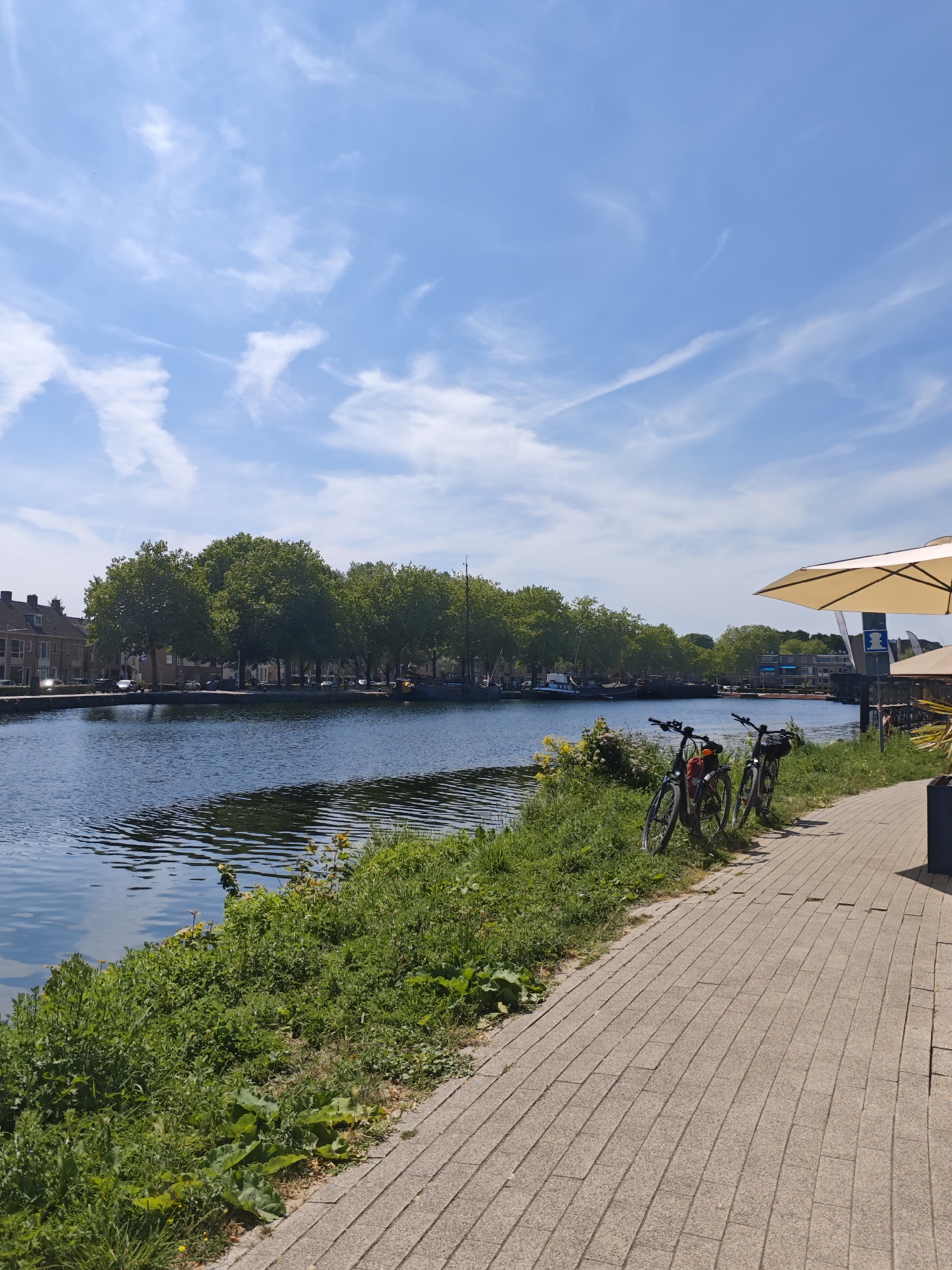 The image depicts a canal scene in the city of Tilburg, Netherlands, on a bright, sunny day. The canal runs horizontally across the frame, reflecting the blue sky and scattered white clouds. The left bank of the canal is lined with residential buildings and a row of mature trees. The right bank features a paved walkway with two bicycles parked alongside a patch of lush green grass and yellow wildflowers. Further along the walkway, a large, beige umbrella provides shade. Several boats are moored on the canal, and a street with cars is visible on the left bank. The overall atmosphere is tranquil and picturesque, suggesting a leisurely day by the water.