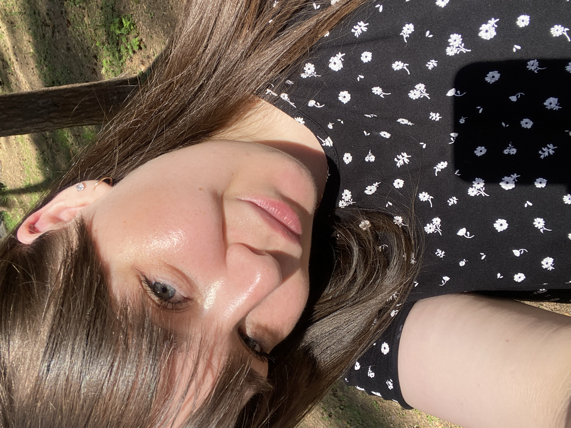 A woman is lying in an outdoor environment, likely a park or wooded area, with her head turned toward the camera. She has light skin, brown hair, and is wearing a black dress with small white floral patterns. The lighting suggests it is daytime, with sunlight casting shadows on her face and clothing. The background shows glimpses of greenery and a tree trunk. There is no information available to locate the image in Votkinsk, Russia.