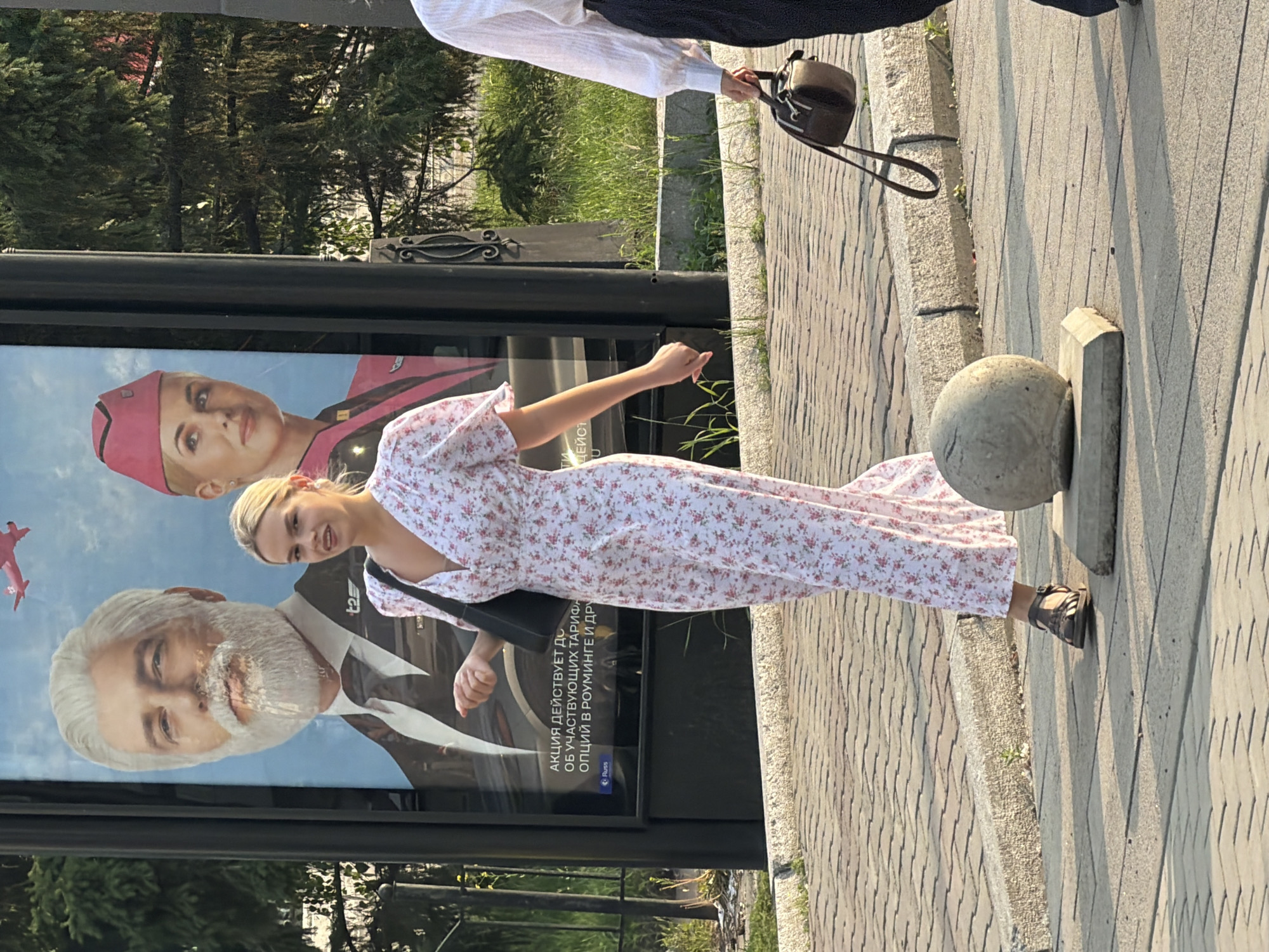 The image shows a young blonde woman in a floral print dress and sandals standing on a sidewalk next to a large advertisement. She is looking slightly off-camera with a neutral expression and has a black handbag slung over her shoulder. The advertisement features portraits of two individuals: a woman in a pink hat and uniform and a man with a white beard. Text is visible on the advertisement in Russian, including "АКЦИЯ ДЕЙСТВУЕТ ДО" (Promotion valid until) and other words related to tariffs and roaming. The setting appears to be an urban environment, possibly a street or public square, with trees and a paved walkway visible. A concrete bollard is also present near the woman's feet. The lighting suggests it is daytime, likely late afternoon due to the long shadows cast on the sidewalk. The scene is shot from a slightly tilted perspective.