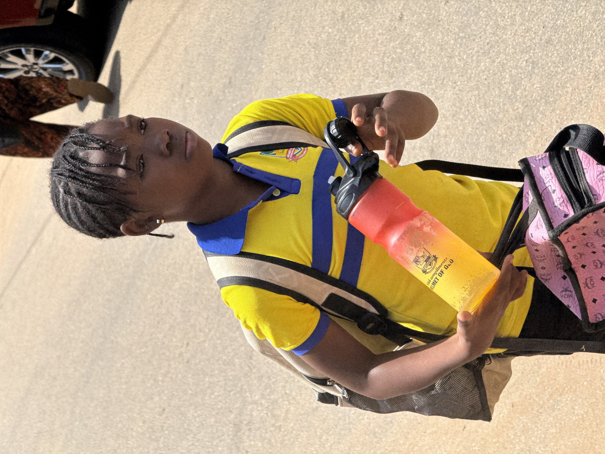 The image shows a young Black person wearing a yellow and blue collared shirt and a backpack. The person is holding a red and yellow water bottle. A pink backpack with a repeating "MCM" logo is also visible. In the background, a car tire and part of a red vehicle can be seen, along with a light-colored, textured ground that suggests an outdoor setting. The lighting indicates it is daytime, with shadows cast on the ground. The person's expression is neutral. There is text visible on the water bottle that reads "RICH COMPLIMENTS. HELMET OF GLO." The scene appears to be in Abuja, Nigeria.
