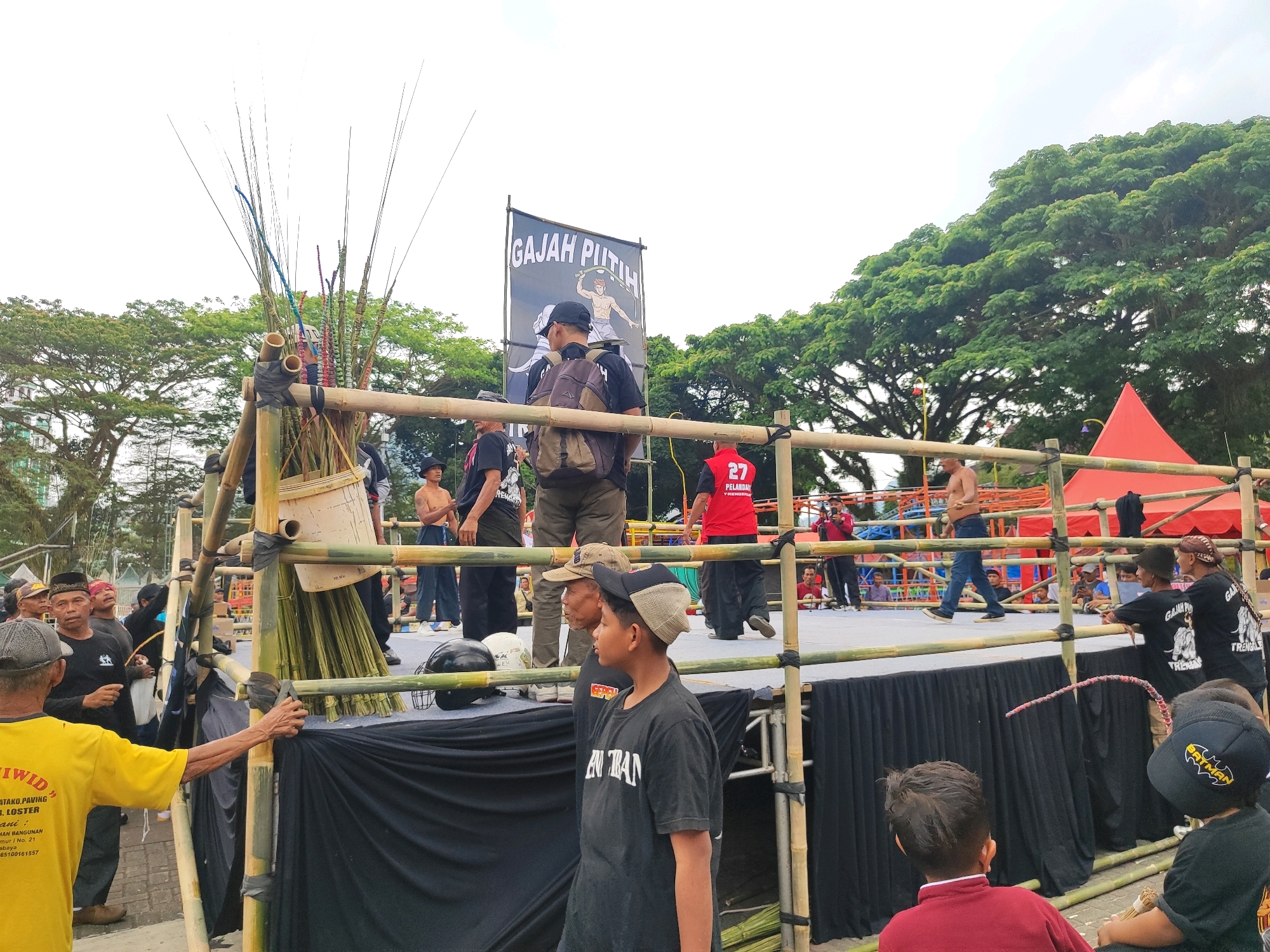 The image depicts an outdoor event, likely a martial arts demonstration or competition, taking place in Trenggalek, Indonesia. A bamboo-constructed boxing ring is the central focus, draped in black fabric at its base. A banner with the text "GAJAH PUTIH" hangs prominently above the ring. Several individuals, mostly men and some boys, are present around and inside the ring. Some are wearing traditional attire, while others are in casual clothing. One person in the foreground is wearing a yellow t-shirt with text related to construction. A red tent is visible in the background, as are lush green trees, suggesting a park or open field setting. The sky is overcast.