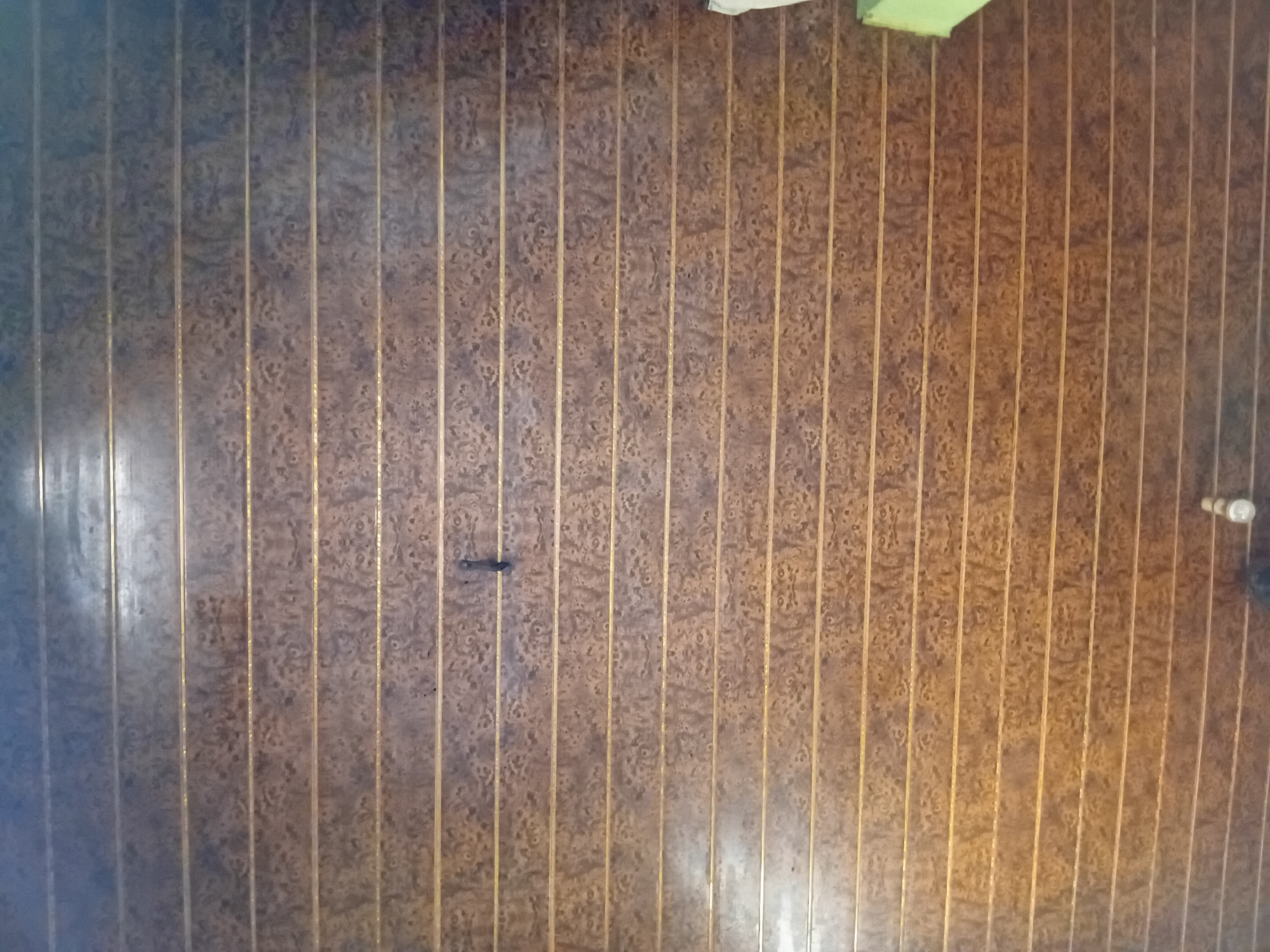 The image shows a close-up view of a wood-patterned ceiling, likely made of vinyl or similar material, with vertical seams. The pattern is a marbled or burl wood effect in shades of brown and gold. The lighting is uneven, with a brighter, cooler light source on the left side, suggesting it may be from a window or artificial light. There is a dark, irregular mark on one of the panels in the upper left quadrant, possibly a stain or scuff. In the upper right corner, a light fixture with visible bulbs can be seen. The ceiling appears to be in an interior space. The context of Nkpor, Nigeria is not visually derivable from the image itself.