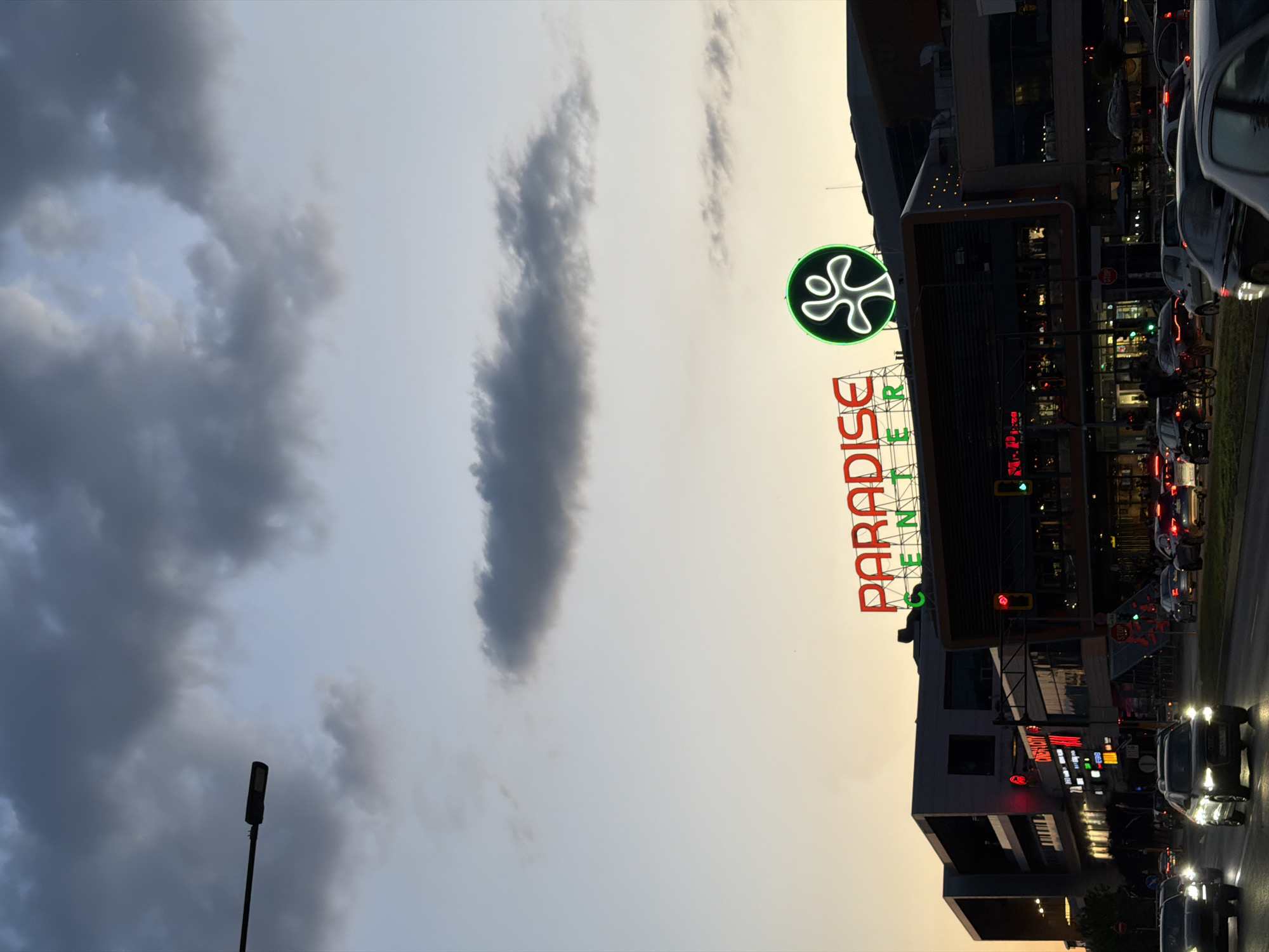 The image shows the Paradise Center shopping mall in Sofia, Bulgaria. The sky is filled with dark clouds, giving the impression of an approaching storm or dusk. The shopping mall's signage is prominently displayed, with the "PARADISE CENTER" name in red neon lights and a circular green neon symbol above it. Cars are lined up on the street in front of the mall, suggesting a busy shopping time. A lone lamppost is also visible against the backdrop of the cloudy sky.