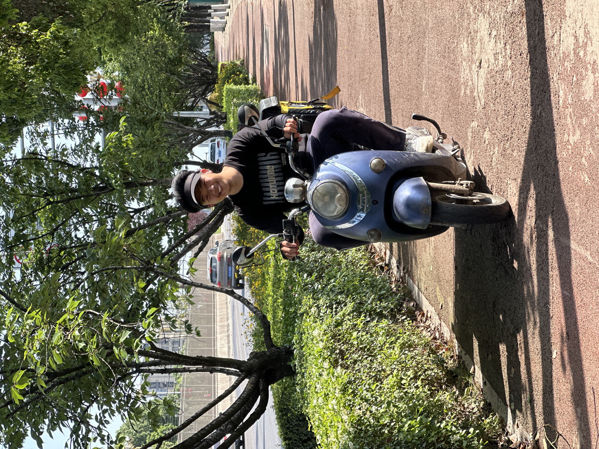 A young Asian man is seated on a blue scooter on a bright, sunny day. He is smiling broadly and looking towards the camera, holding the handlebars. He wears a black short-sleeved t-shirt with white text that reads "HotHunter," dark pants, a black visor cap, and white sneakers. A yellow delivery bag or backpack is visible on the back of the scooter.
The scene takes place outdoors on a paved, reddish-brown pathway, likely a sidewalk or park path. Lush green trees with dense foliage line the path, creating shade and casting long, distinct shadows across the ground and a textured wall, indicating clear weather and bright daylight, possibly mid-morning or afternoon. Green bushes also border the path. In the background, partially obscured by the trees, a road or street with a silver car can be seen. Several red lanterns are discernible hanging amongst the trees in the distance, suggesting a cultural context consistent with Jiangjin, China.