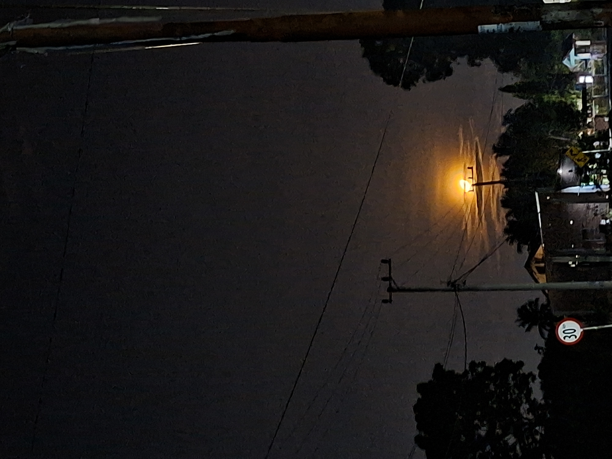 The image shows a night scene in Kutaglumpang, Indonesia. A street lamp illuminates the area, casting a bright orange glow. Power lines crisscross the sky. A speed limit sign reading "30" is visible on the right. There are utility poles, trees, and silhouettes of buildings. The sky is dark and overcast. The perspective is tilted, with the foreground dominated by darkness.