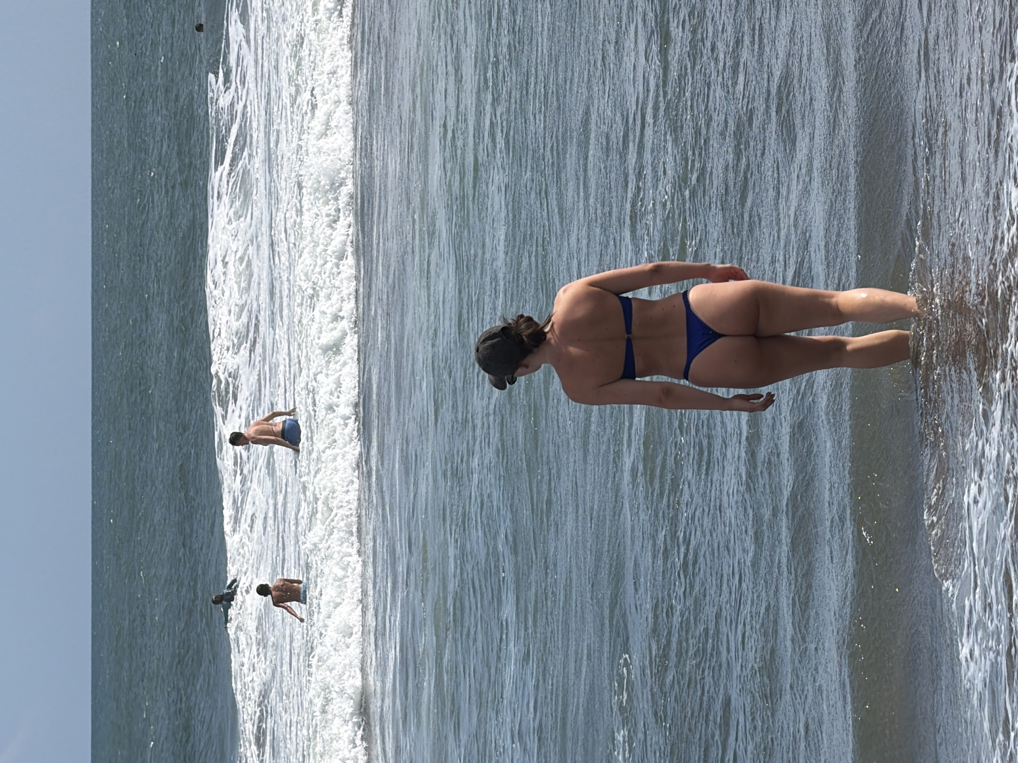 In the city of North Kuta, Indonesia, a woman in a blue bikini and a dark baseball cap stands with her back to the viewer, wading into the ocean. She is positioned in the foreground of the image. Further out in the water, in the middle ground, several other individuals are visible. One person is partially submerged, with only their head visible above the waves. Two other individuals, appearing to be surfers, are in the ocean, one on a surfboard and the other without. The background consists of the vast expanse of the ocean under a bright, clear sky. The water is a deep blue, with white waves crashing and forming foam closer to the shore.