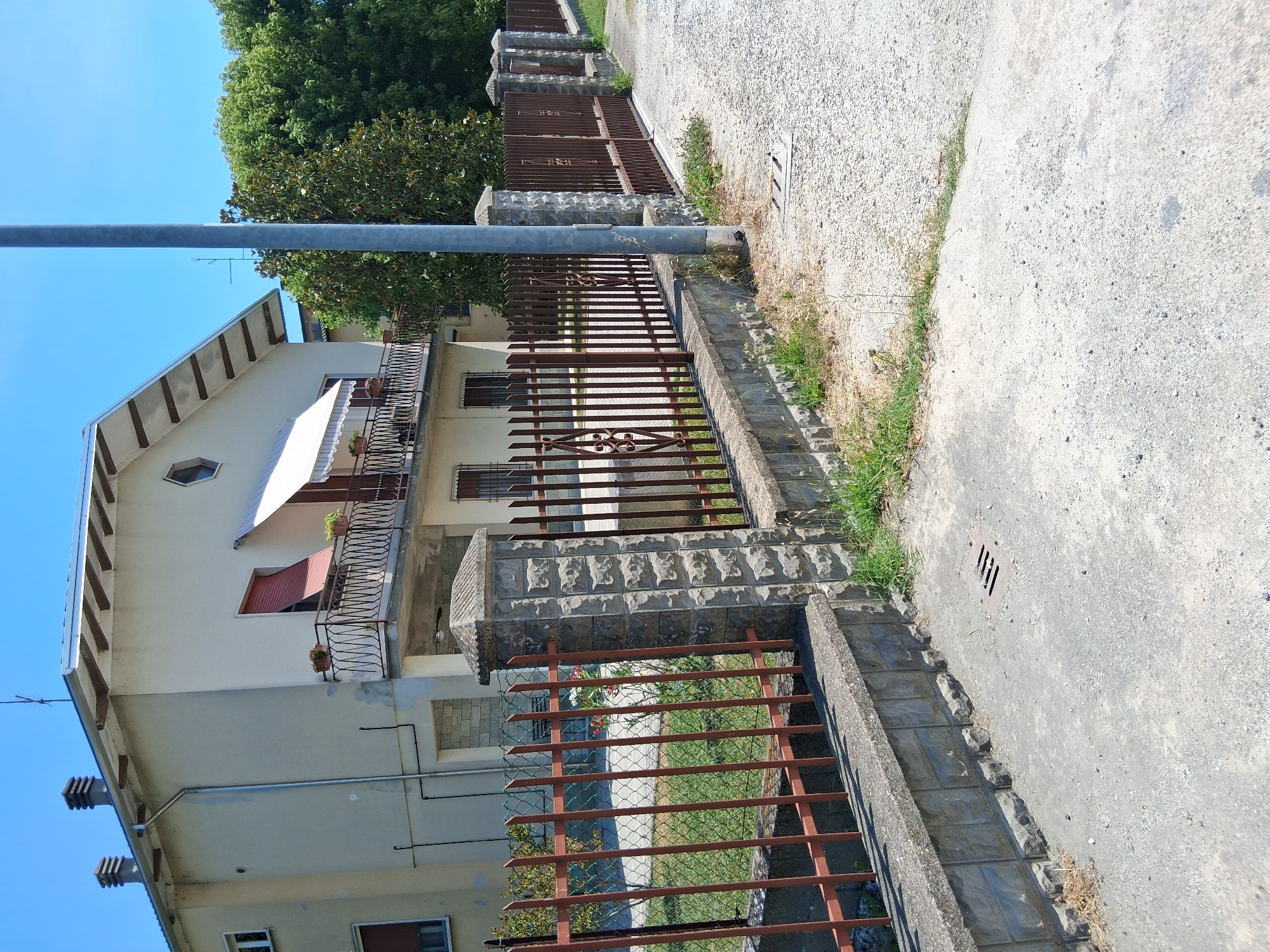 The image depicts a street scene in Colorno, Italy, under a clear blue sky. In the background, a two-story house with a light-colored facade and a sloping roof is visible. The house has several windows, some with dark shutters, and a balcony with a black metal railing adorned with potted plants. An awning extends over the balcony. A fence with vertical brown metal bars supported by stone pillars lines the property. The fence is set on a low concrete wall. Beyond the fence, a patch of green grass is visible. In the foreground, a grey paved area, possibly a sidewalk or driveway, shows some gravel and grass growing at the edges. A metal grate, likely a drain cover, is embedded in the pavement. A tall grey metal pole, possibly a street light or utility pole, stands near the fence. The overall environment suggests a residential area.