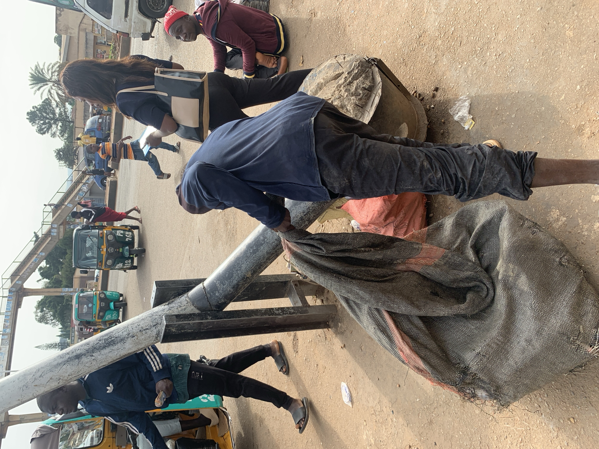 In Gura Top, Nigeria, a man in a blue long-sleeved shirt and dark pants is attending to a large piece of burlap sacking draped over a metal structure. He appears to be adjusting or gathering the material. Nearby, another individual in a burgundy hooded jacket and red beanie is seated on the ground, looking towards the left. The scene unfolds on a dusty, outdoor surface, with a large metal pole prominent in the foreground, partially obscured by the burlap. In the background, several auto-rickshaws are visible, along with pedestrians and a pedestrian bridge overhead, suggesting a busy public area. The overall impression is one of everyday activity in a bustling Nigerian locale.