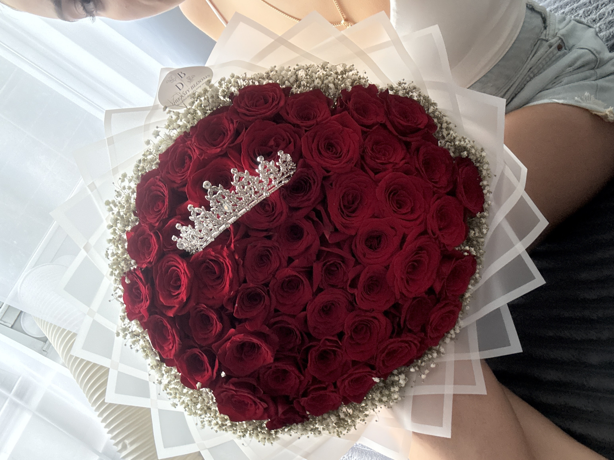 An overhead shot shows a bouquet of deep red roses accented with baby's breath and topped with a delicate silver tiara. The bouquet is wrapped in translucent white paper with geometric patterns and is held by a person whose lap and legs are visible. The person is wearing a white top and light blue denim shorts. The background consists of white curtains and blinds, suggesting an indoor setting. A tag with the logo "DB DREAMY BLOOMS" is attached to the bouquet. A portion of the person's face, including an eye and eyebrow, is visible in the upper right corner of the frame. The image is sharp and well-lit. The specific location in New York City, United States of America, is not identifiable from the visual information provided.