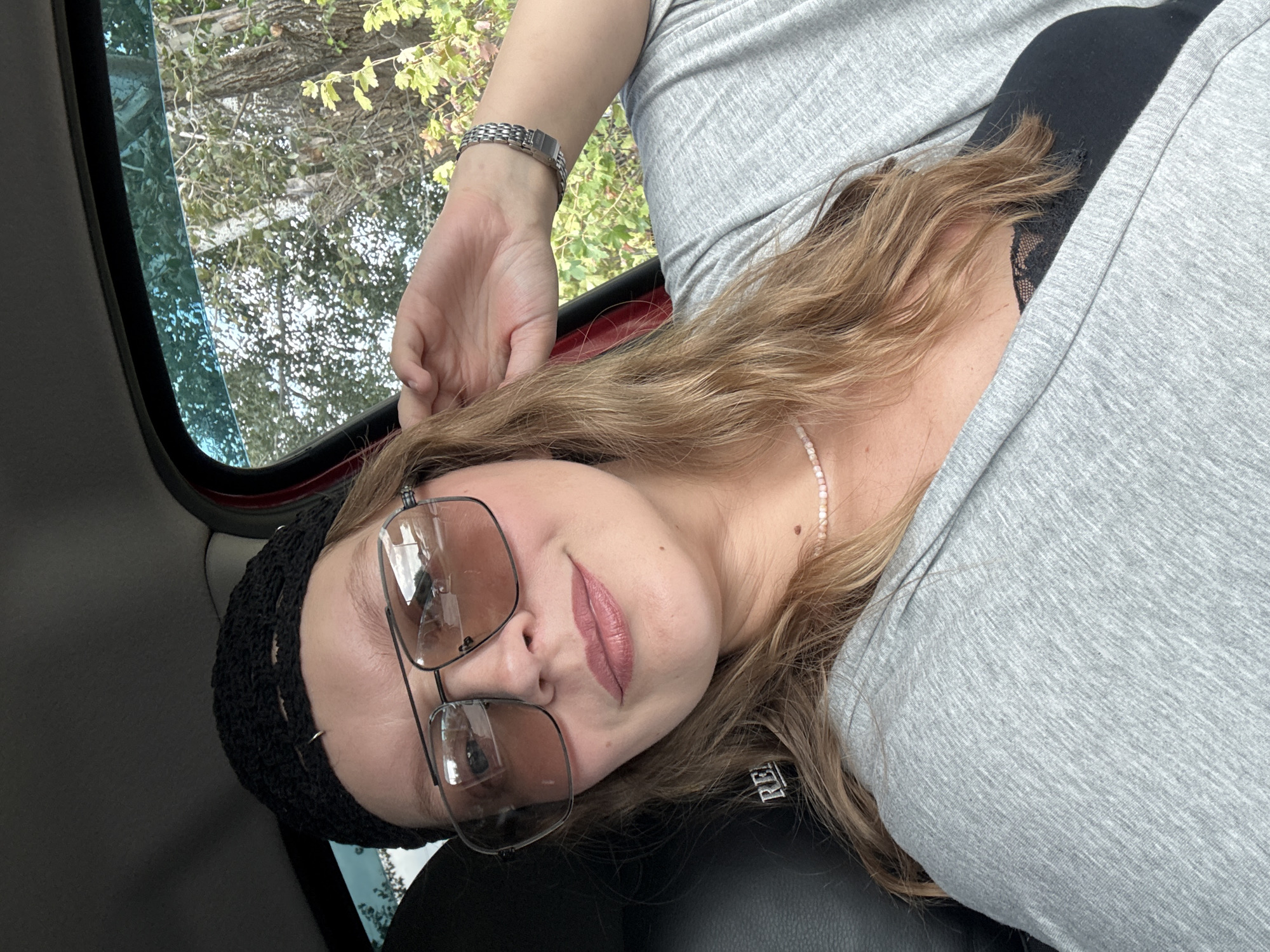 A woman is sitting in the driver's seat of a car, possibly in Aksay, Kazakhstan. She is wearing a black crochet beanie, large aviator-style sunglasses with brown lenses, and a light grey cardigan over a black top with lace detailing. A thin pink beaded necklace is visible around her neck. Her right hand is raised, touching her hair near her ear. Her hair is medium-length and light brown. The background visible through the car window shows trees and foliage, suggesting an outdoor setting. The car's interior, including the black leather seat and part of the dashboard, is also visible.