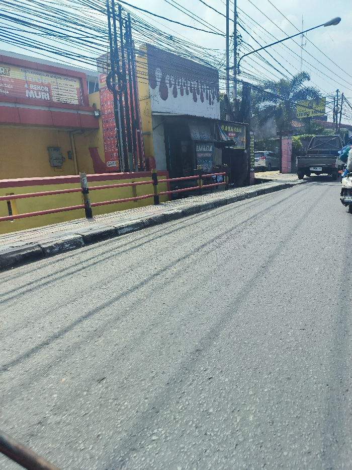 The image shows a street in Cileunyi, Indonesia, at latitude -6.933 and longitude 107.724. The street is paved with asphalt and has a red and yellow guardrail on the right side. There are several power lines running overhead. On the right side of the street, there is a building with a sign that says "BAKARAK" and another sign with unreadable text. In front of the building, there is a truck parked. The truck is facing away from the camera. There are other vehicles visible in the distance. There is a motorcycle parked in the foreground. The image is taken from a low angle and shows the street from a perspective of a person looking down at the street. The sky is clear and there is a light breeze. The street is deserted. There are no people in the image. The asphalt is grey and has visible tire marks.