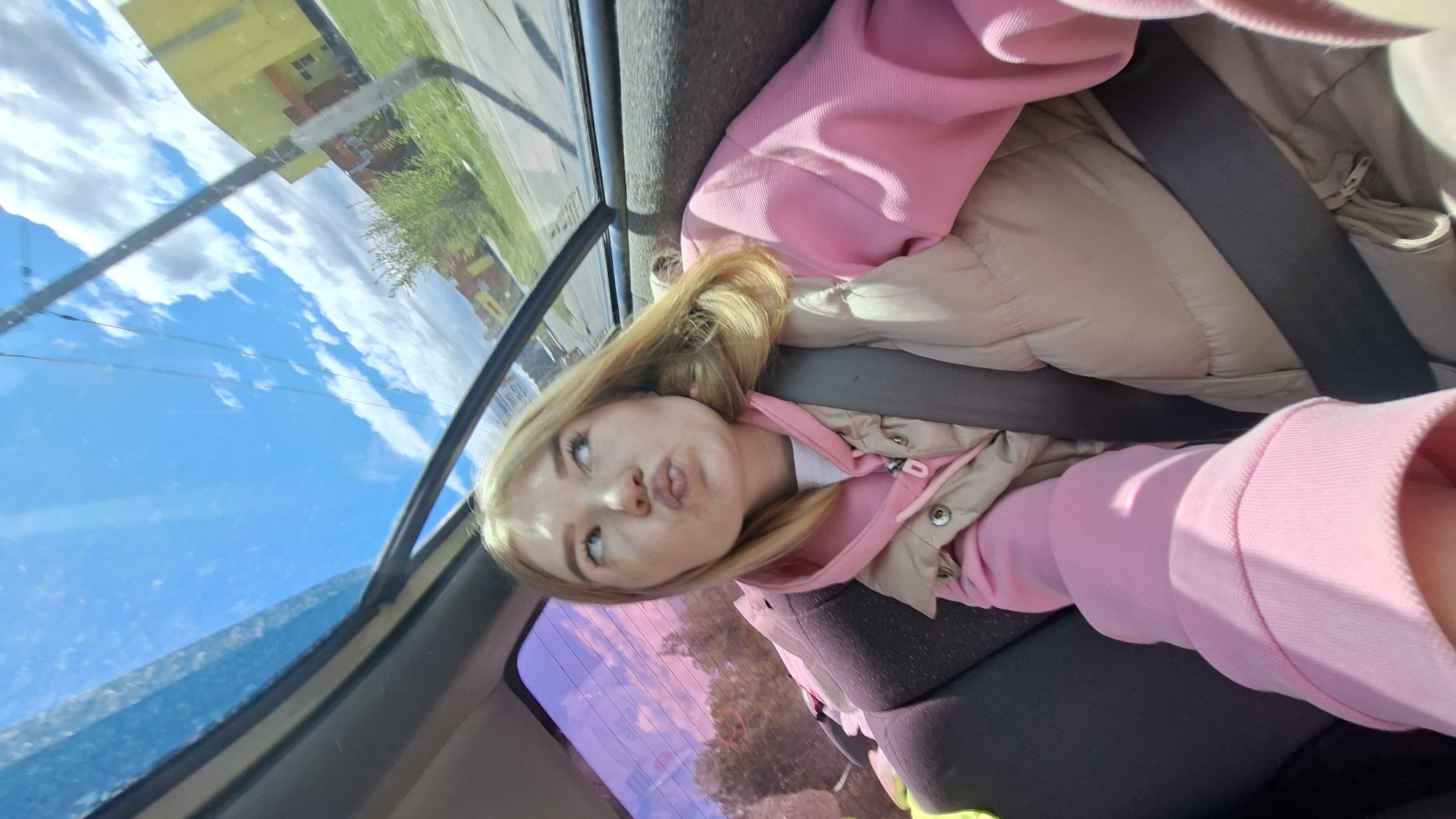 A young woman with blonde hair is seated in the back of a car, wearing a pink hoodie and a tan-colored jacket. She is wearing a seatbelt and has a playful expression on her face. The window shows a reflection of the sky with clouds and a glimpse of a yellow building with greenery, potentially situated in Chaikovsky, Russia. The car's interior includes grey seats and a dark headliner.
