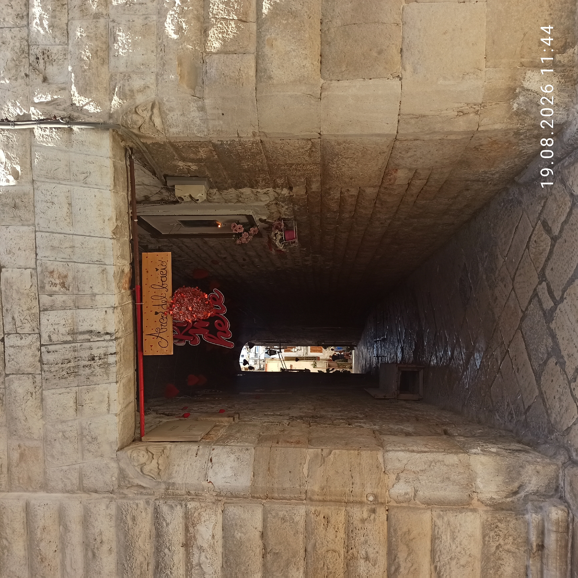 This image captures a narrow, stone-paved alleyway in Ruvo di Puglia, Italy, under a stone archway. The archway is decorated with red heart-shaped ornaments and a sign that reads "Arco del bacio" (Arch of the kiss). A circular, red, textured object hangs beneath the sign. The walls of the alley are constructed from large, rough-hewn stones, with some showing signs of wear and discoloration.
To the left of the archway, embedded in the stone, is a small, carved relief of a winged figure with a cherubic face. Further down the alley, two figures, appearing to be men, are visible walking away from the camera towards the open end of the alley. The alley opens to a brighter area with buildings and what appear to be lampposts.
On the right side of the alley, a window is visible with a planter box containing pink flowers. A small electrical box is mounted on the exterior wall to the left of the archway. The paving stones in the alleyway are uneven and have a wet or reflective sheen, suggesting they may be damp or polished. The overall impression is of an old, historic part of the city. A timestamp at the bottom right of the image indicates "19.08.2026 11:44".