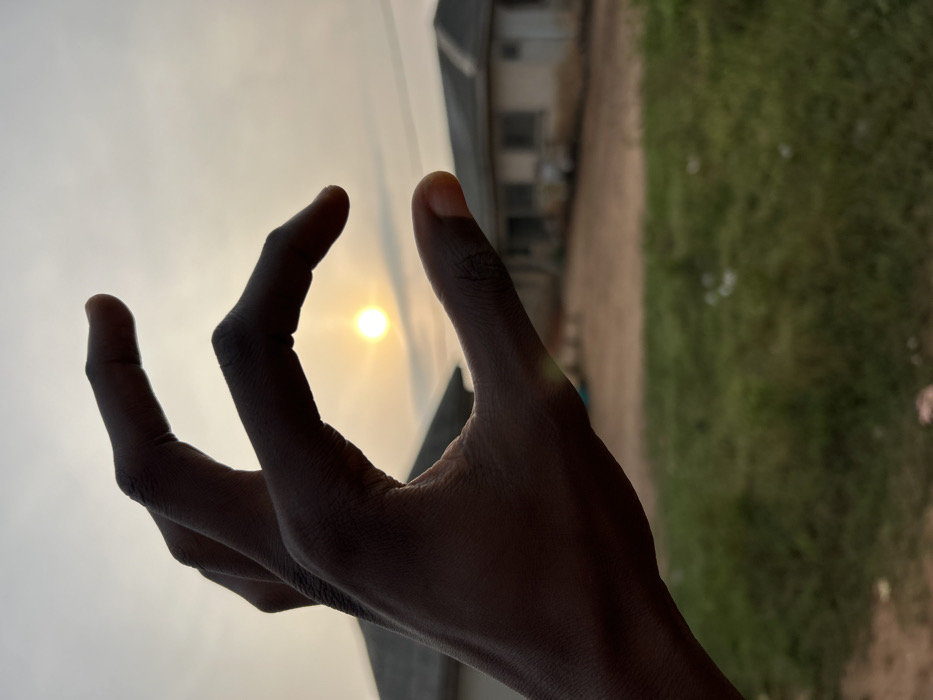 A hand reaches toward the sky, fingers curved to cradle the sun. Silhouetted against the bright sky, the hand is dark. Background elements include part of a building and greenery. The scene suggests a connection between humanity and nature, with the hand attempting to capture or hold the sun.
