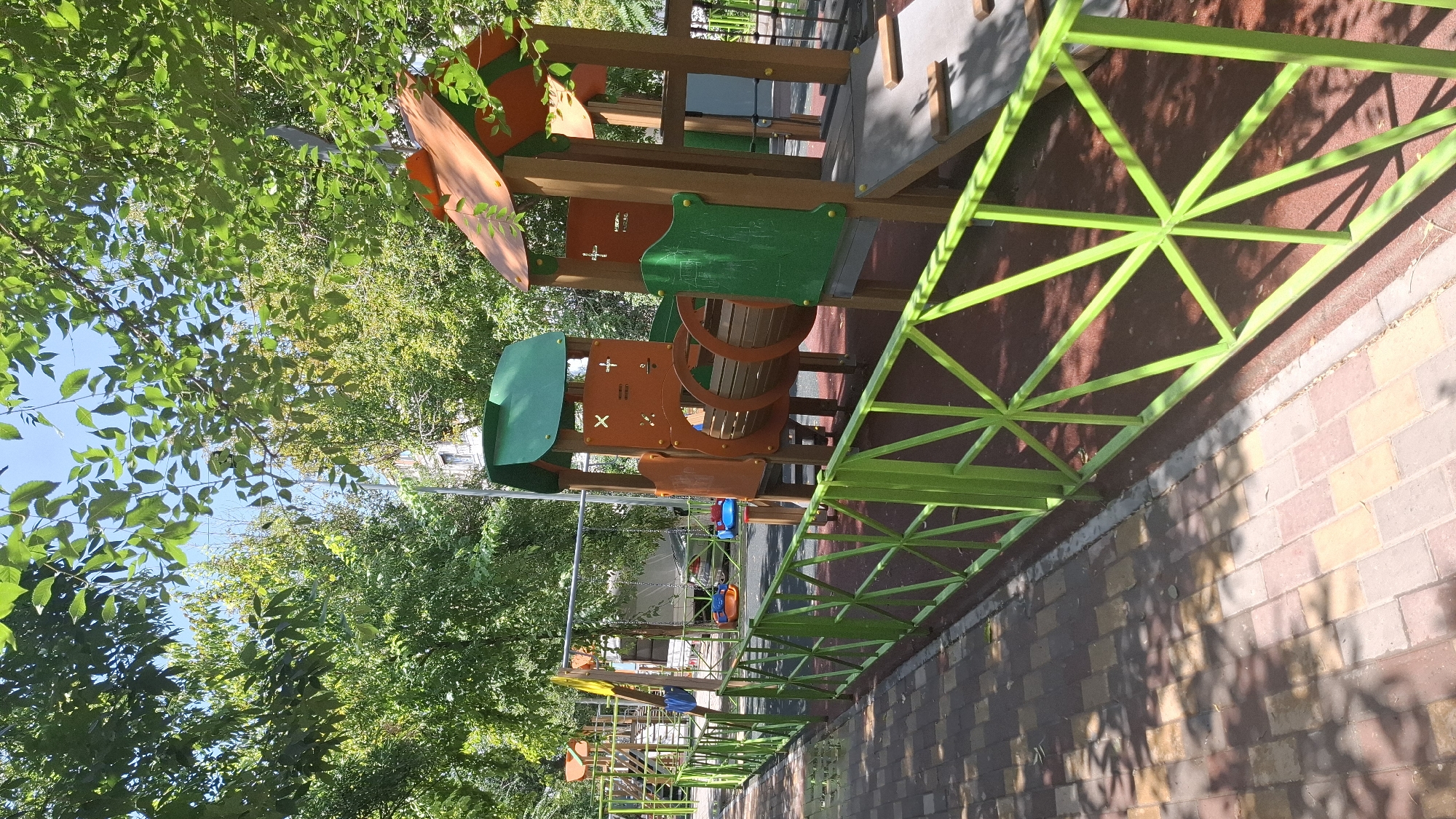 The image depicts a brightly colored playground situated outdoors, likely in a residential area of Shymkent, Kazakhstan. The playground features wooden play structures with orange and green accents, including a tunnel and a slide. A bright green metal fence with a geometric pattern encloses the playground area. The ground within the enclosure appears to be a dark red safety surface, while the pathway leading to the playground is paved with multi-colored rectangular stones. Lush green trees with dense foliage provide shade over the scene, and the sky is visible through the branches. Sunlight casts dappled shadows on the ground. In the background, a multi-story building and a parked car can be seen, suggesting an urban or suburban setting. There are no people visible in the image.